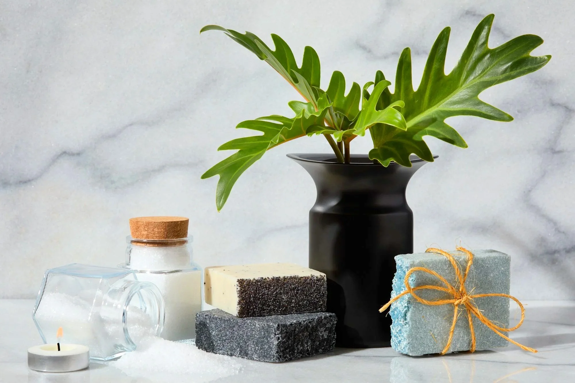 A collection of skincare products and decorative elements on a white surface with a gray stone background. Includes a black vase with green leaves, a small glass jar with a cork lid, layered soap bars, a small tealight candle, and a blue soap tied wi