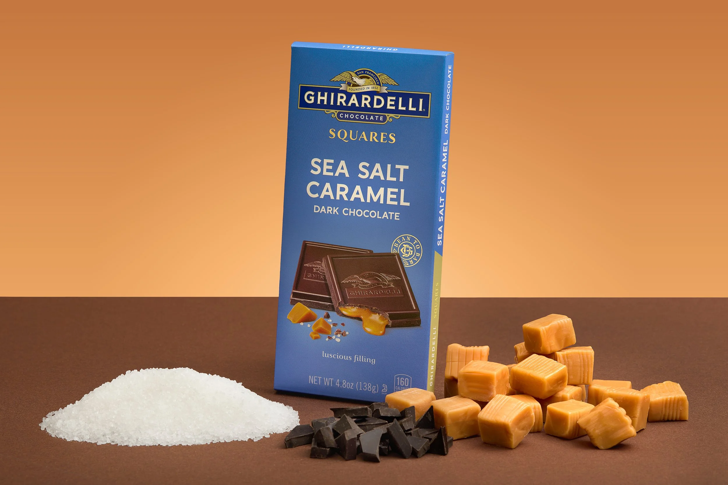 A box of Ghirardelli Sea Salt Caramel dark chocolate squares with caramel and caramel pieces scattered around, along with some white salt and black chocolate chunks, on a brown surface with a brown background.