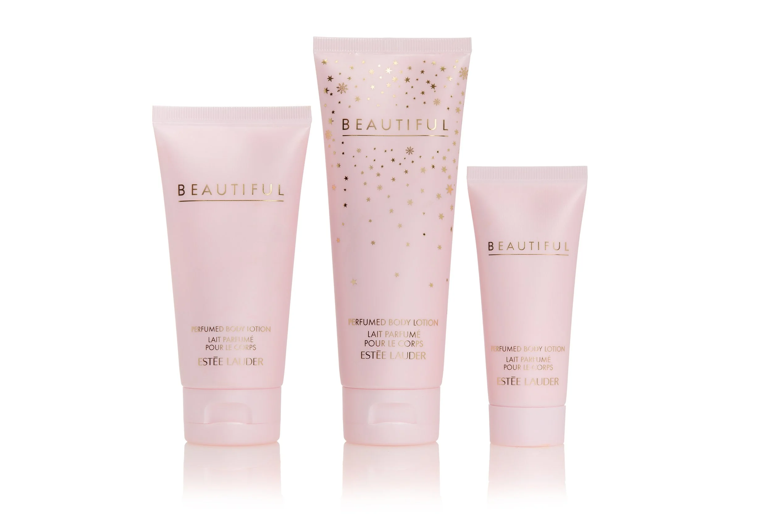 Pack shot of three pink tubes of Estée Lauder Beautiful perfumed body lotion with gold star and snowflake designs, standing upright on a reflective surface.