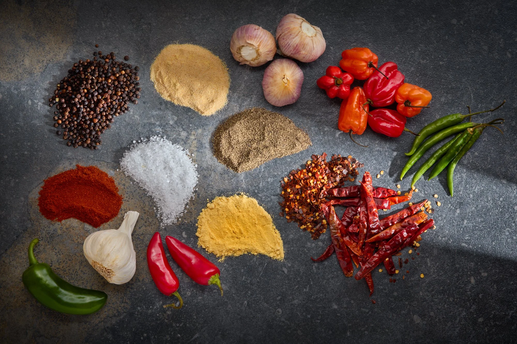 Various spices including black pepper, garlic, dried chili peppers, and colorful fresh peppers on a dark surface.