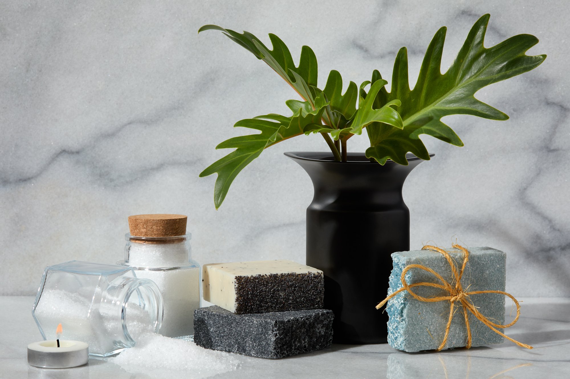 A collection of skincare products and decorative elements on a white surface with a gray stone background. Includes a black vase with green leaves, a small glass jar with a cork lid, layered soap bars, a small tealight candle, and a blue soap tied wi
