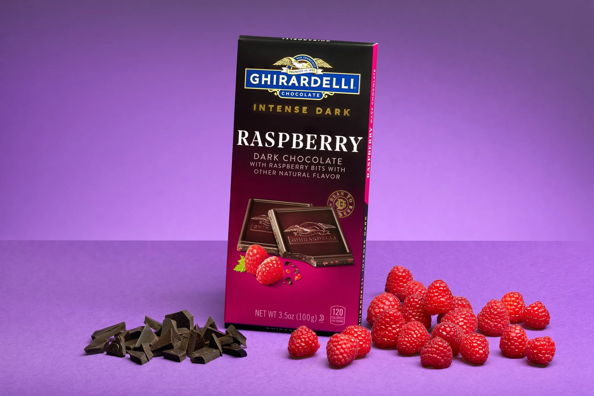 A Ghirardelli dark chocolate bar of raspberry flavor with raspberries, dark chocolate pieces, and a purple background and foreground.