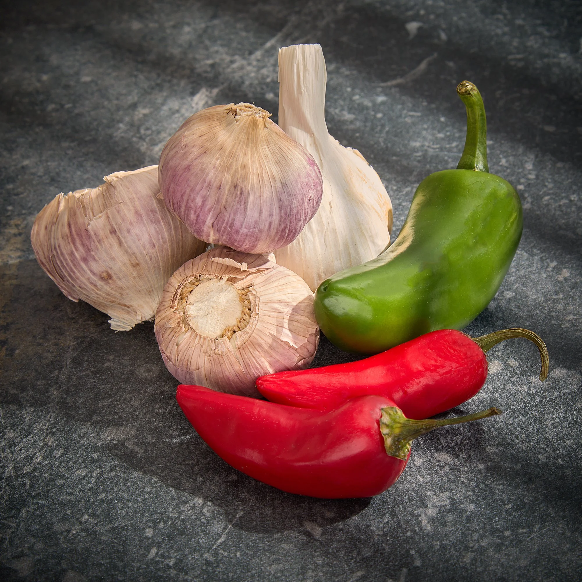 Garlic bulbs, a green bell pepper, and red chili peppers on a dark countertop.