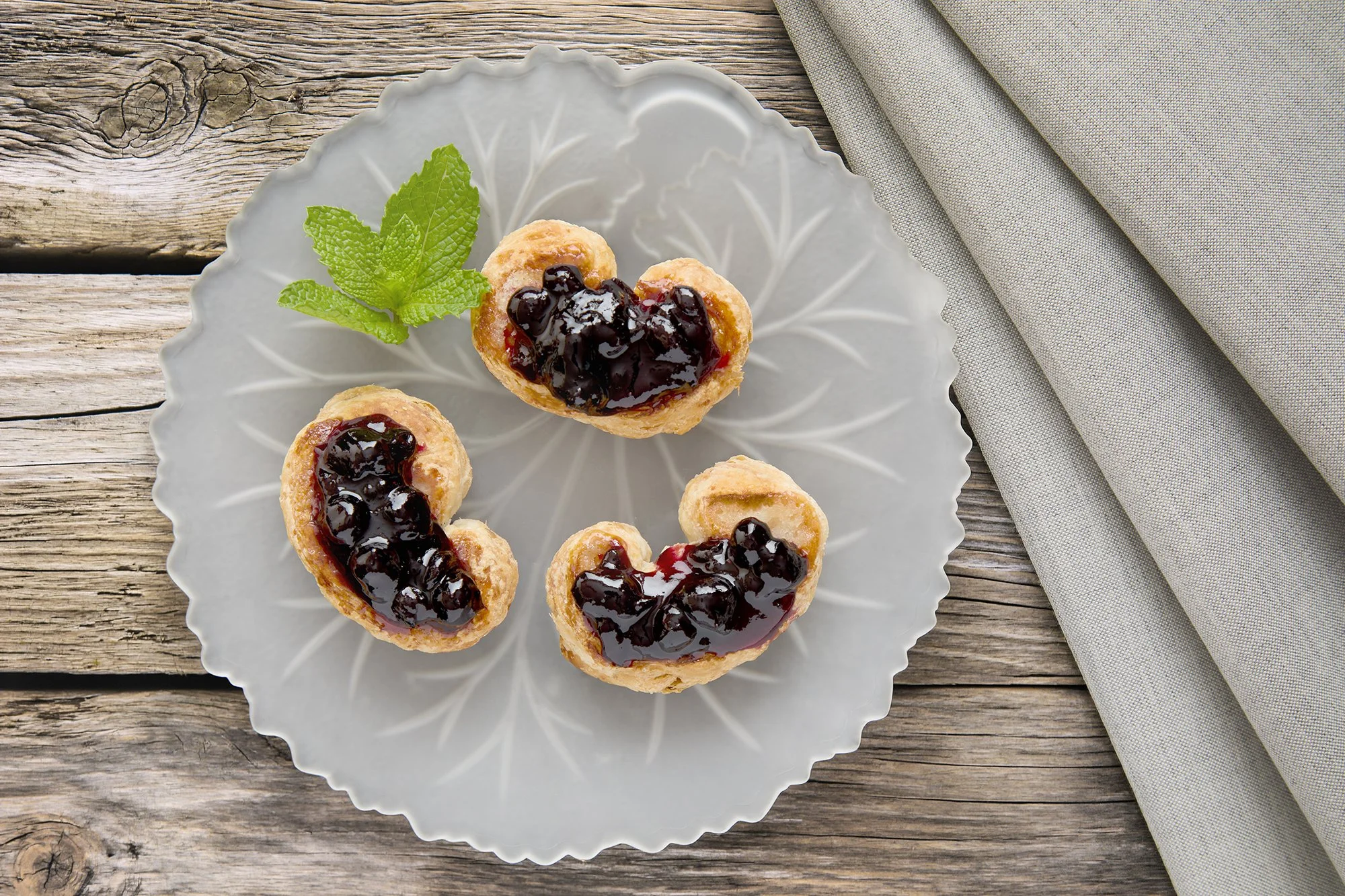 Heart-shaped pastry bites topped with dark blueberry jam arranged on a decorative glass plate with a sprig of fresh mint, placed on a rustic wooden table with a folded beige cloth nearby.