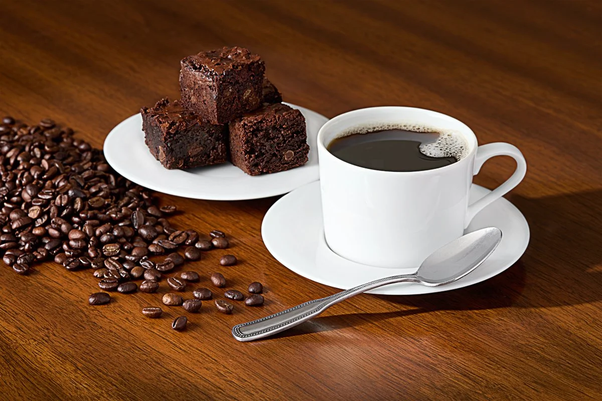 A white cup of black coffee on a saucer with a silver spoon, three chocolate brownies on a white plate, and scattered coffee beans on a wooden table.