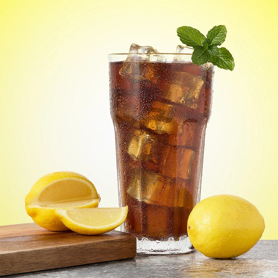 A glass of iced tea with condensation, garnished with a sprig of mint, placed on a surface along with a halved lemon and a whole lemon on yellow gradient background..