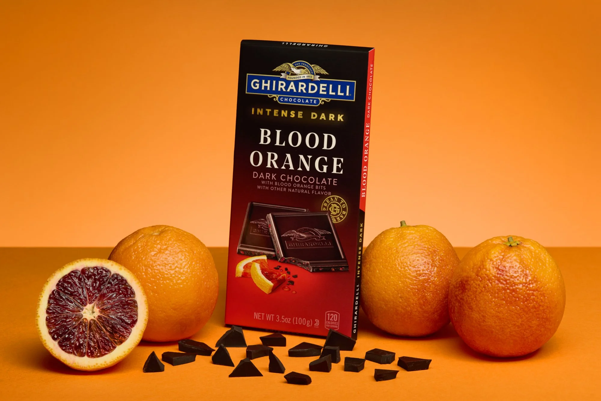 A Ghirardelli intense dark blood orange chocolate bar surrounded by blood oranges, one halved to show its interior, with dark chocolate pieces scattered in front on a orange background.