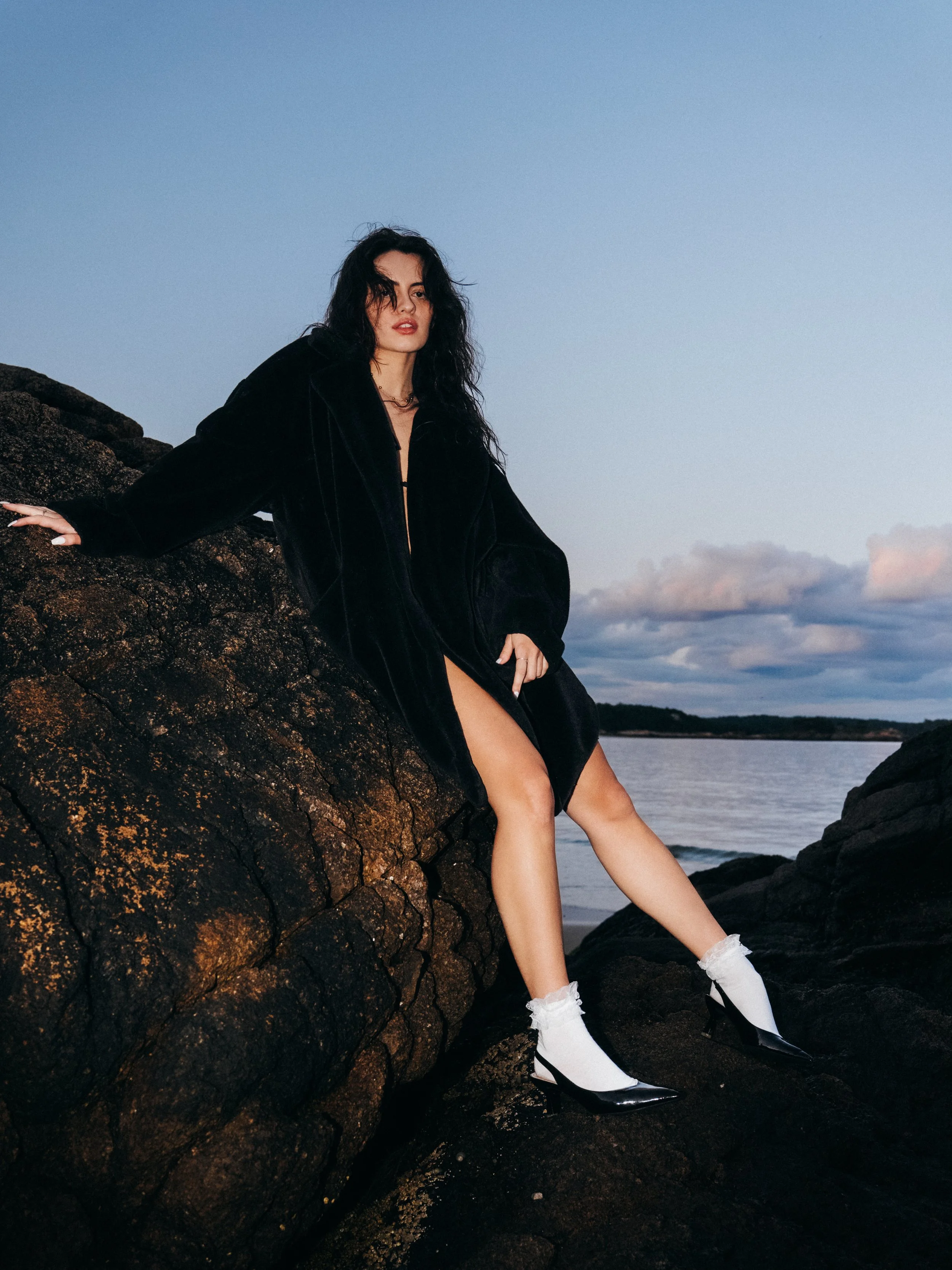 A woman in a black jacket and high heels sitting on rocks near a body of water during sunset, with clouds in the sky.