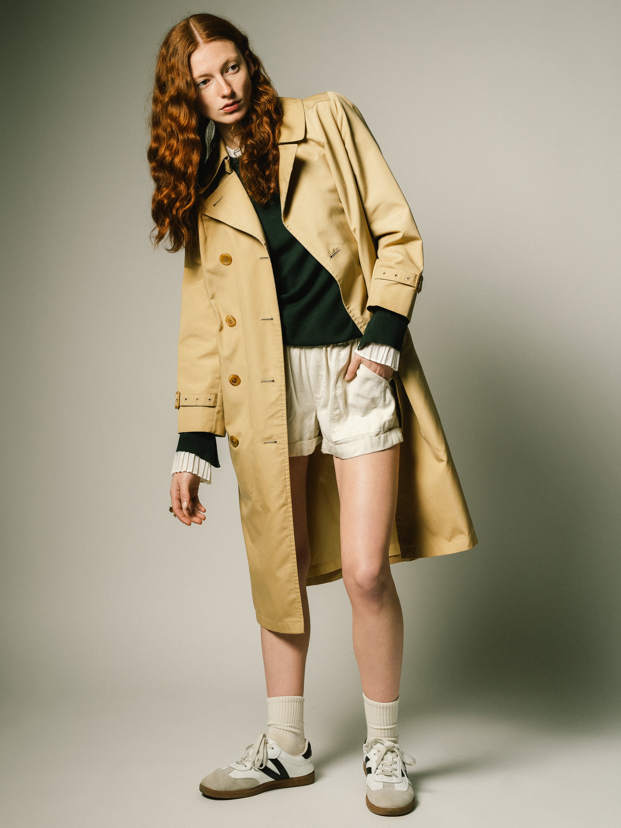 A young woman with long red hair standing against a plain background, wearing a beige trench coat, black sweater, white shorts, white sneakers, and beige socks, with her right hand in her pocket and looking to her left.