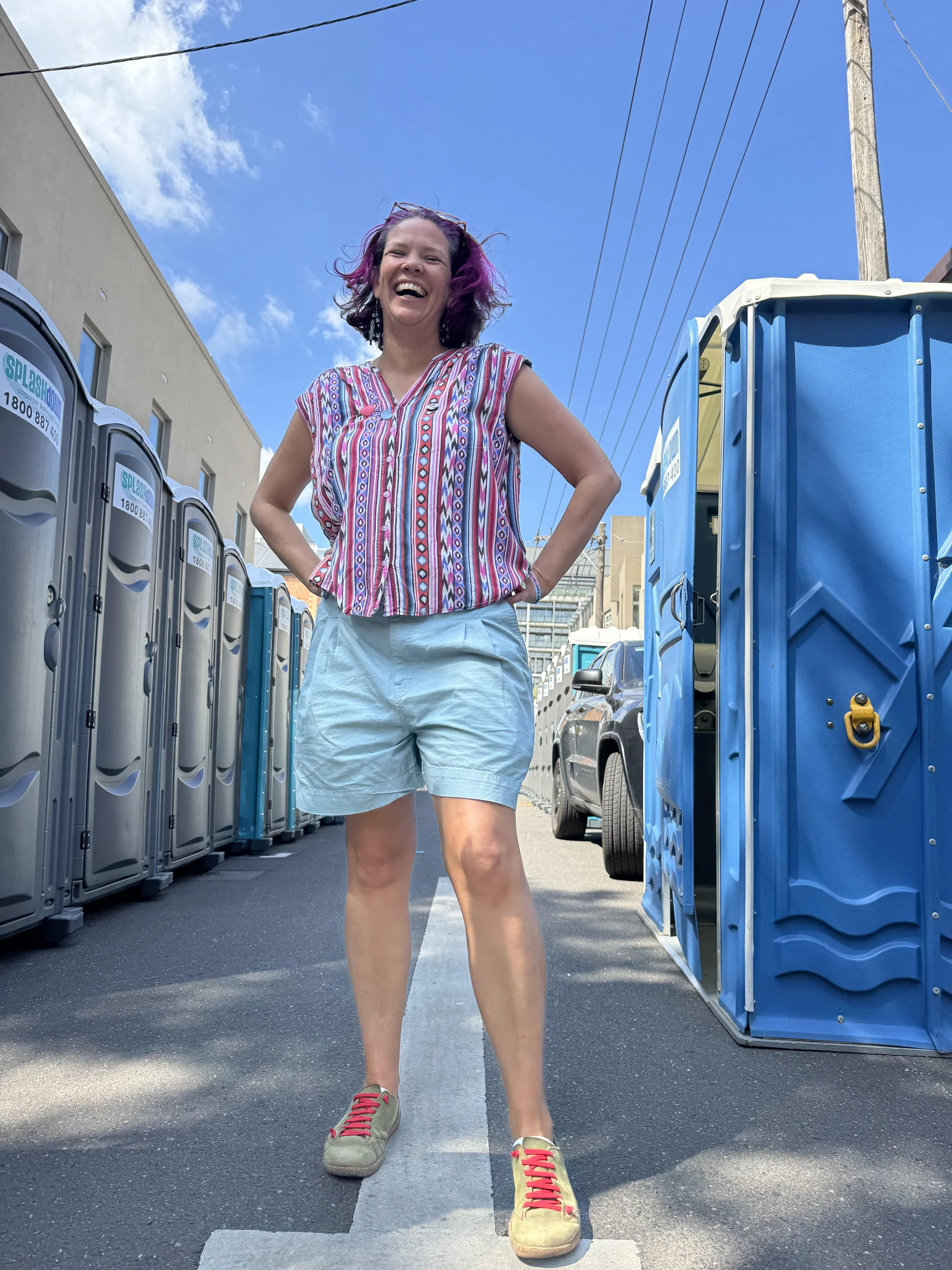 A woman who is smiling standing in a power pose infront of rows of portable toilets.