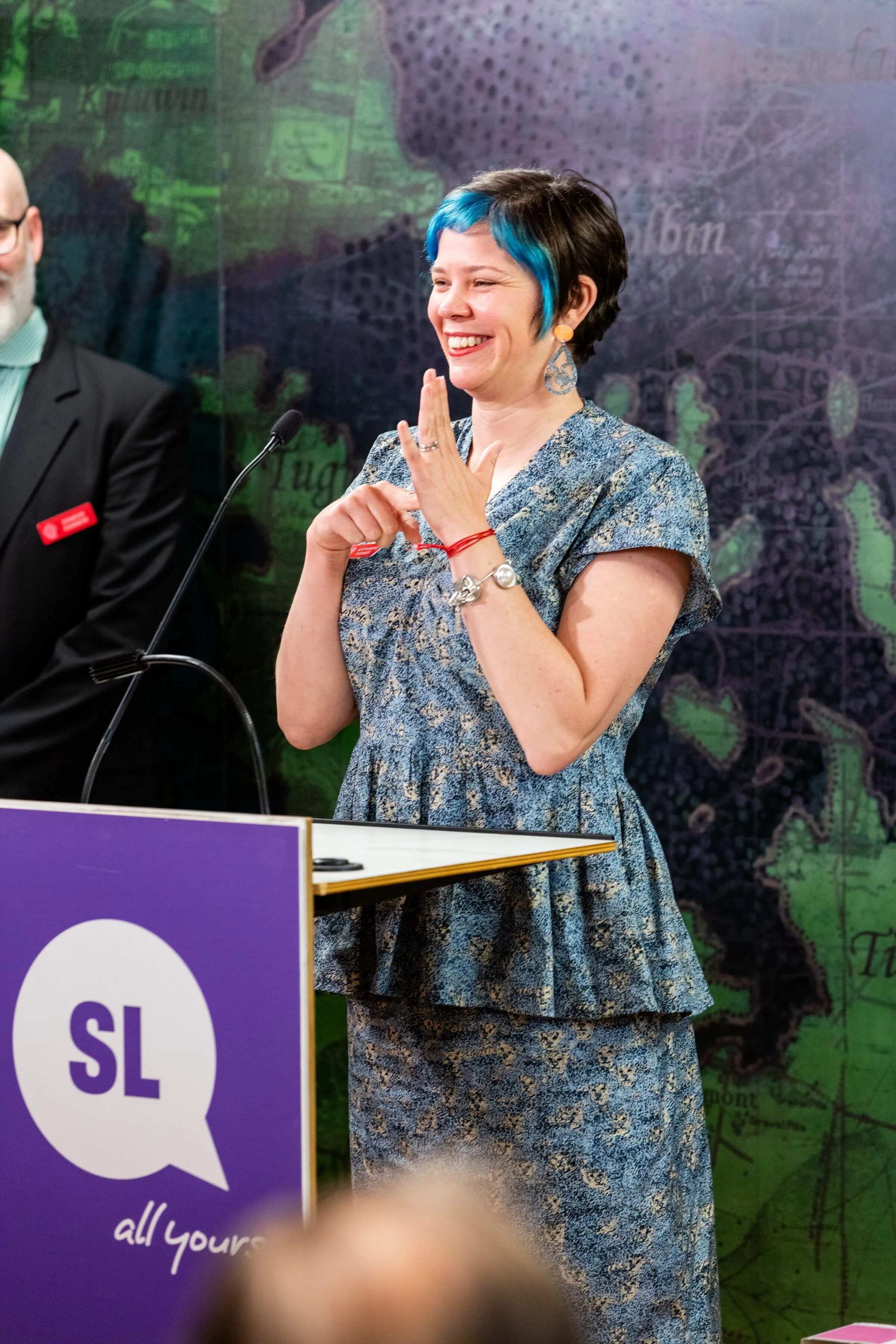 Katherine is at a podium and is signing 'toilet' in Auslan.