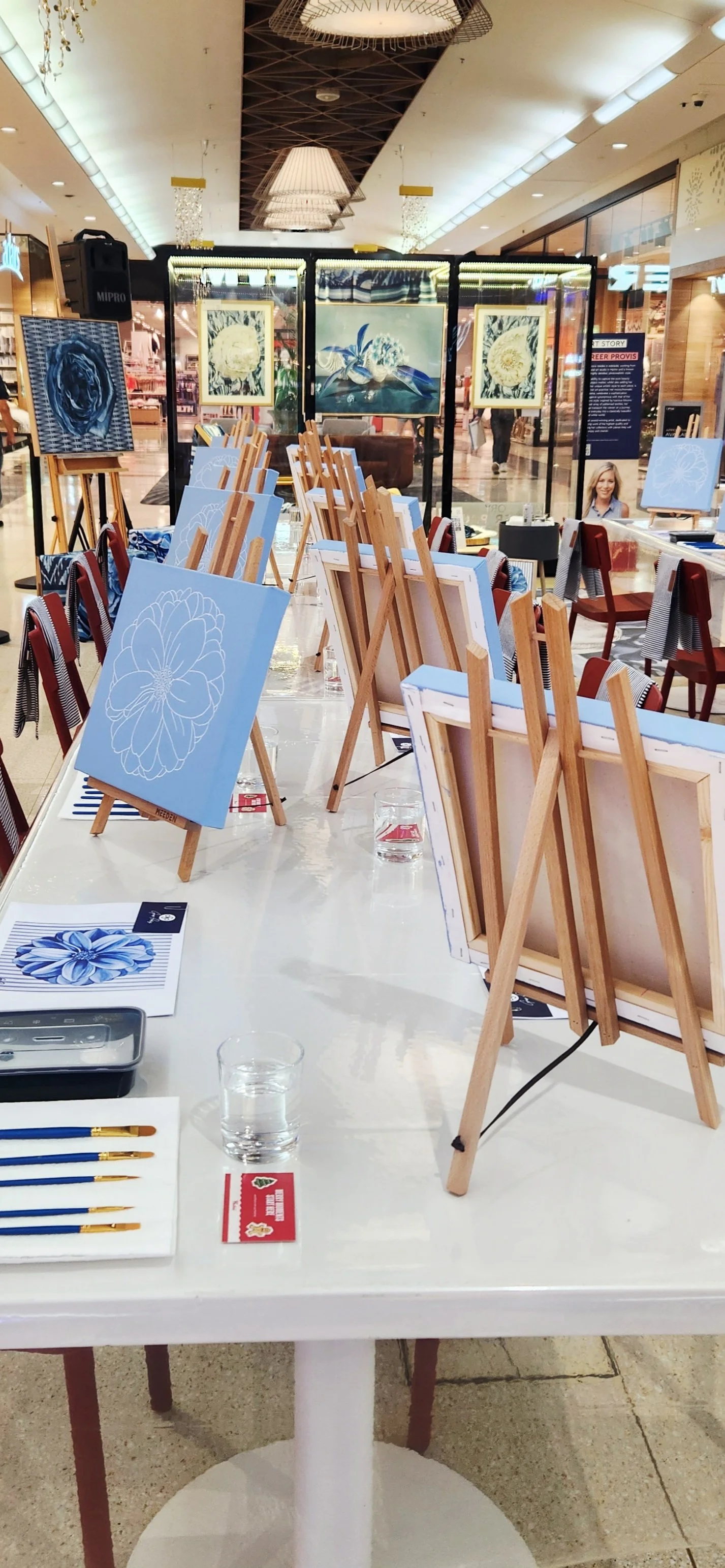 Art exhibit setup for Westfield Marion with canvases on easels featuring blue and white floral designs, a folding screen displaying floral paintings, and tables with art supplies and water glasses. This was Greer Provis' November 2025 workshop.