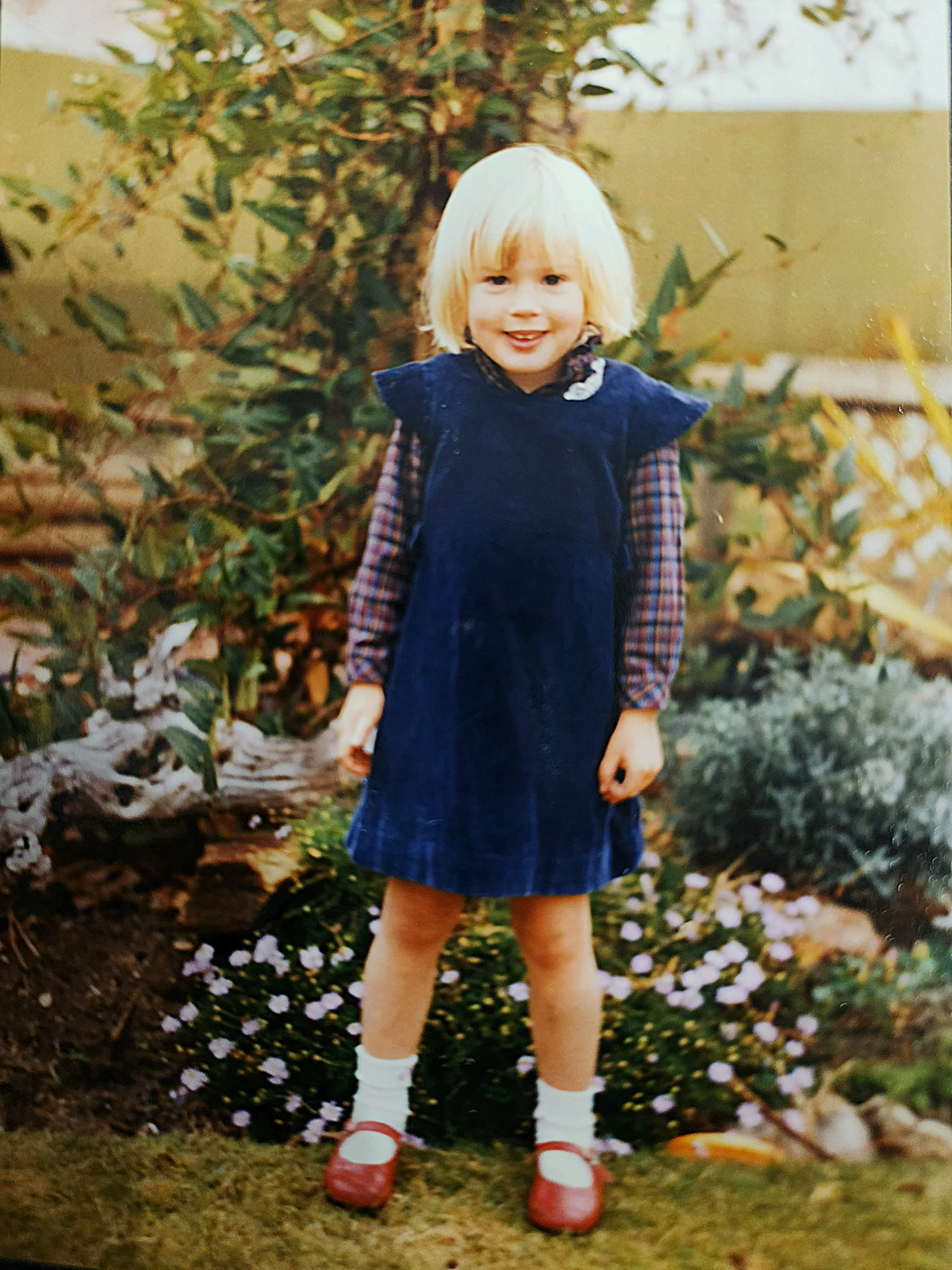 A young girl with blonde hair and fair skin, wearing a navy blue dress, plaid long sleeve shirt, white socks, and red shoes, standing outdoors in front of a garden with flowers and greenery.