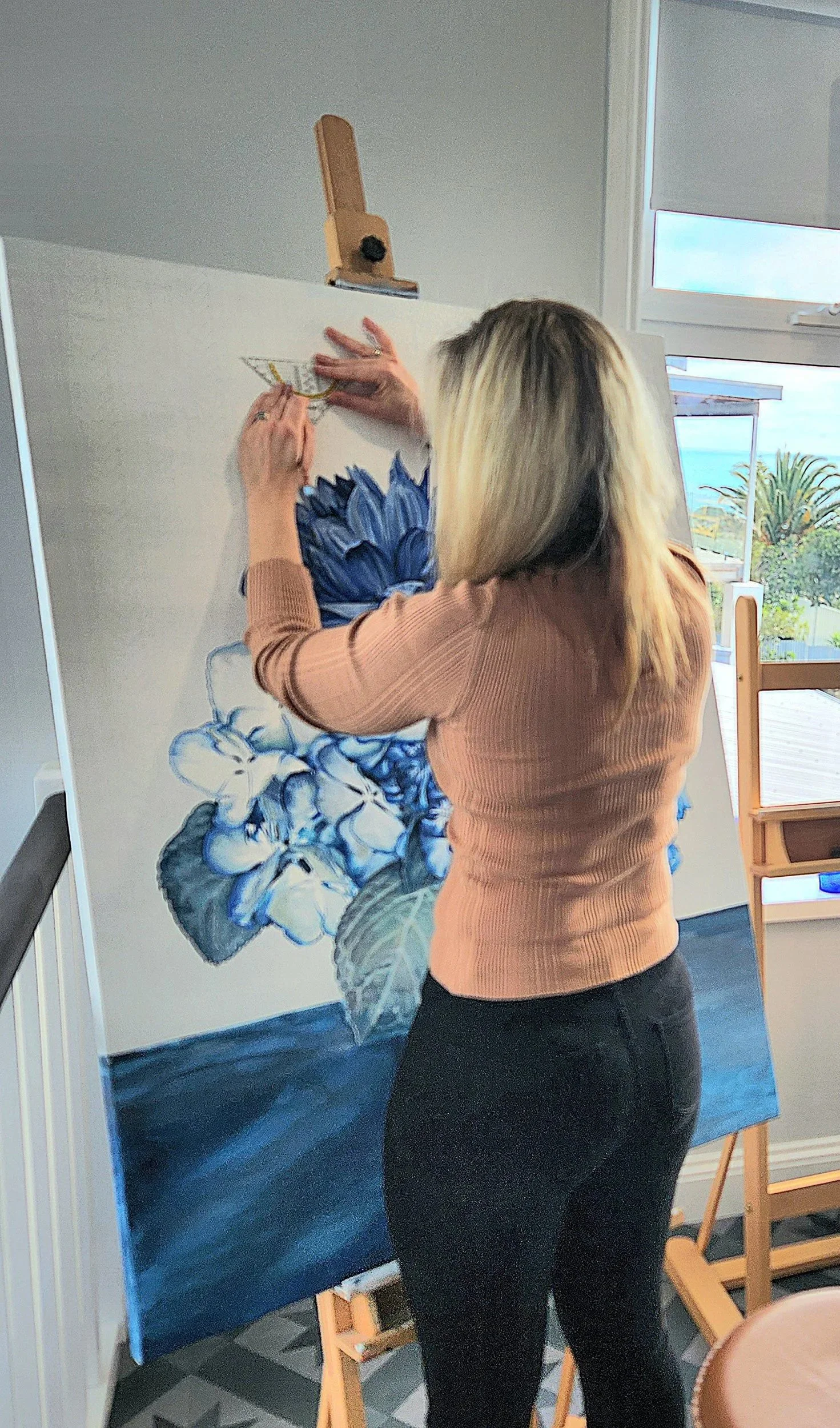 Greer Provis in her studio painting a large floral artwork of blue and white flowers on an easel.