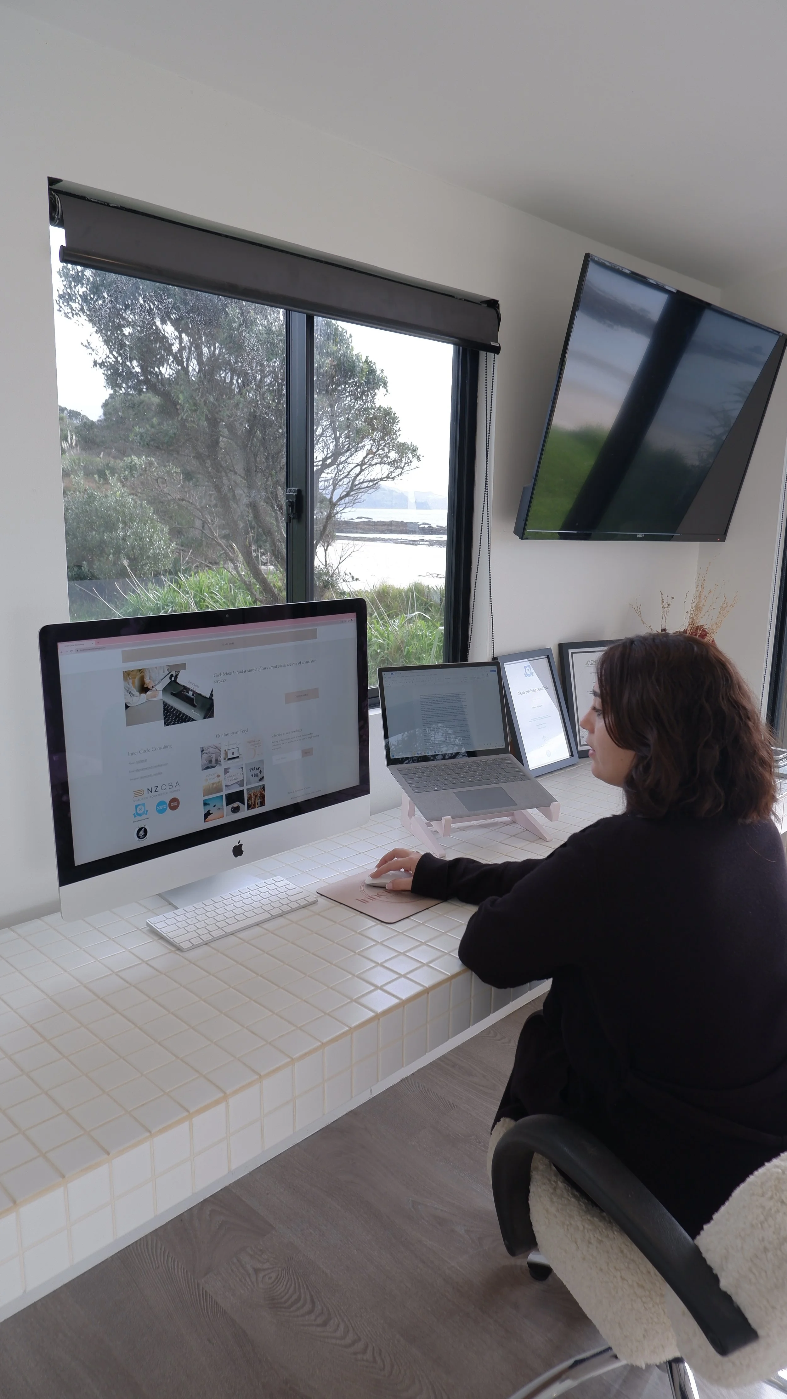 Woman working at a white desk with multiple screens in a bright room with large window showing trees and water outside.