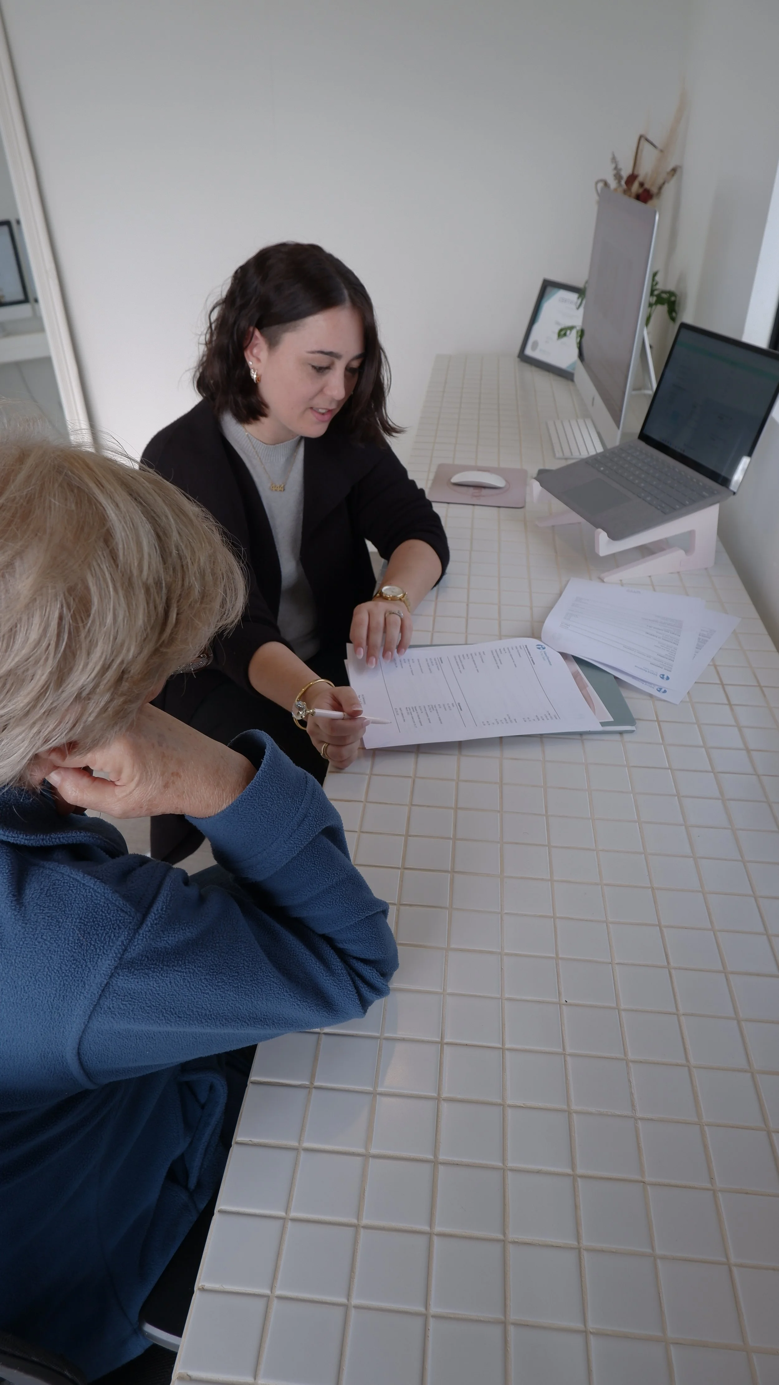A woman and an older person seated at a white tiled table, reviewing documents with a computer monitor and laptop on the desk.