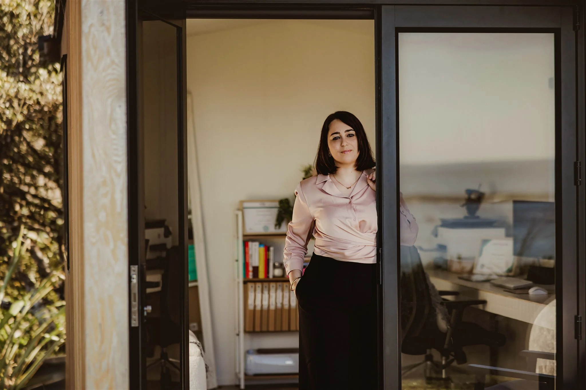 A woman standing at a glass door, wearing a pink shirt and black pants, smiling inside a modern home interior.