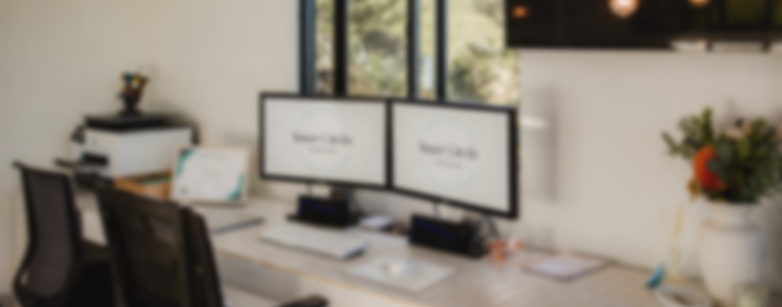 Blurred office workspace with dual computer monitors, a keyboard, and a side vase with flowers.