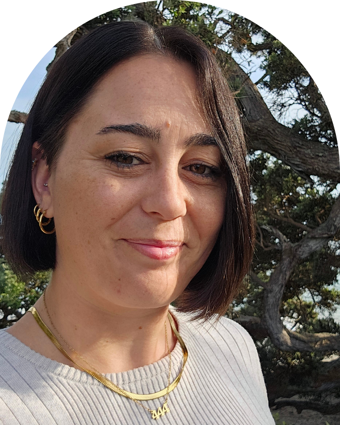 Close-up of a woman with short dark hair, wearing gold jewelry, smiling outdoors with a tree and blue sky in the background.