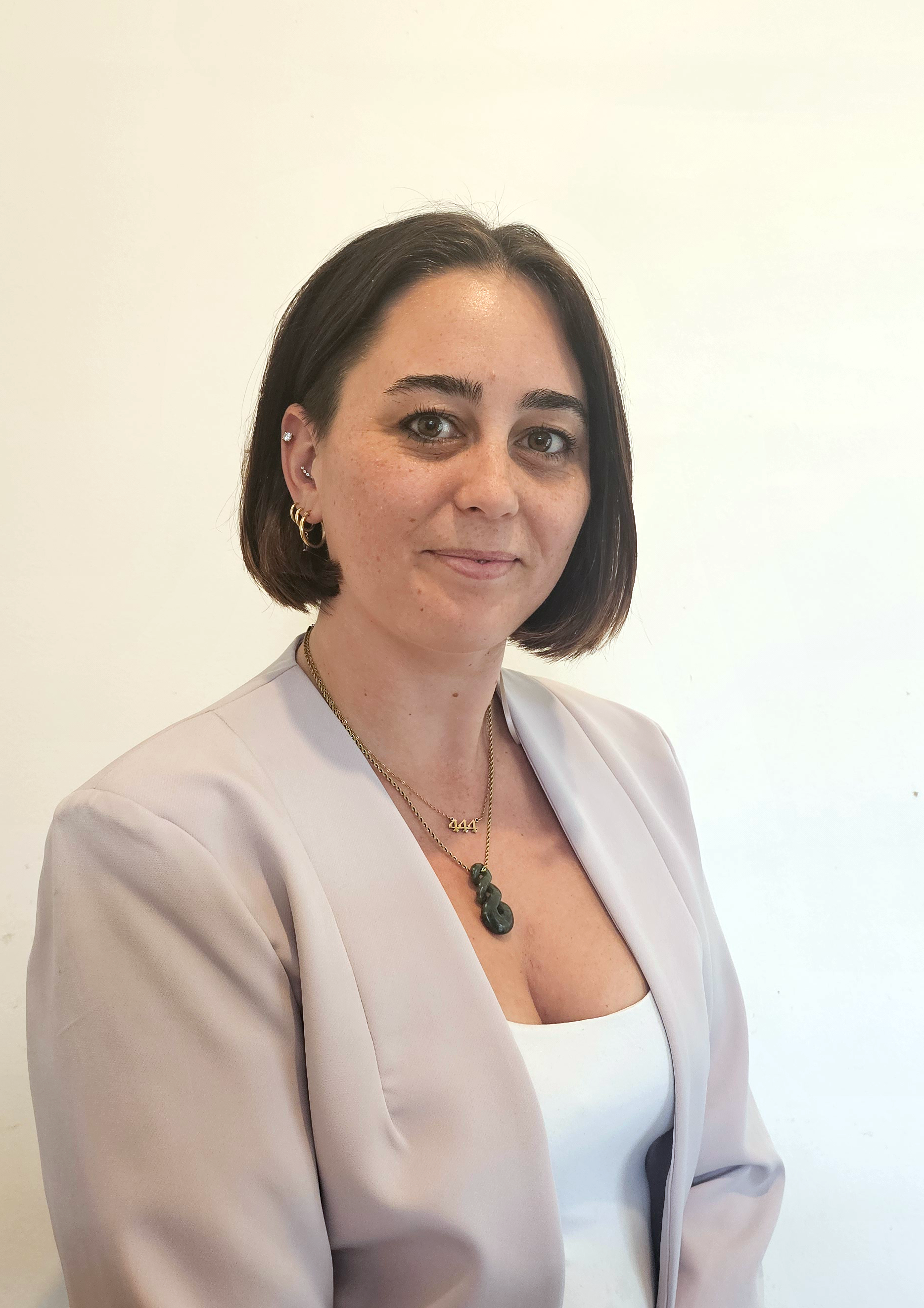A woman with short dark hair, wearing a light beige blazer, white top, and gold jewelry, standing against a plain white background.