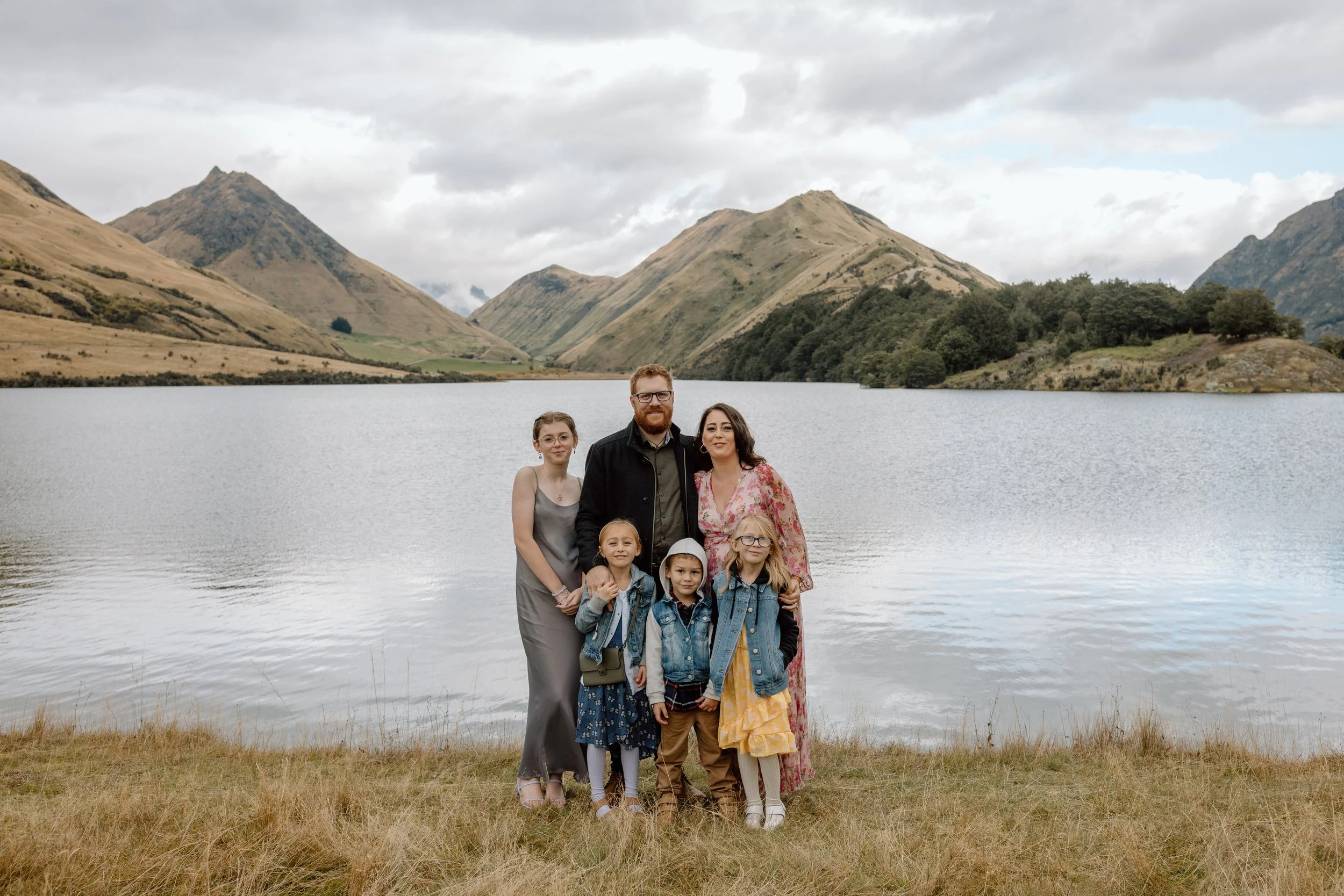 A family of seven standing by a lake with mountains in the background. The adults include a man with glasses and a beard wearing a dark jacket, and a woman in a floral dress. The children include five girls, some wearing denim jackets and one wearing glasses, all smiling and posing for the photo.