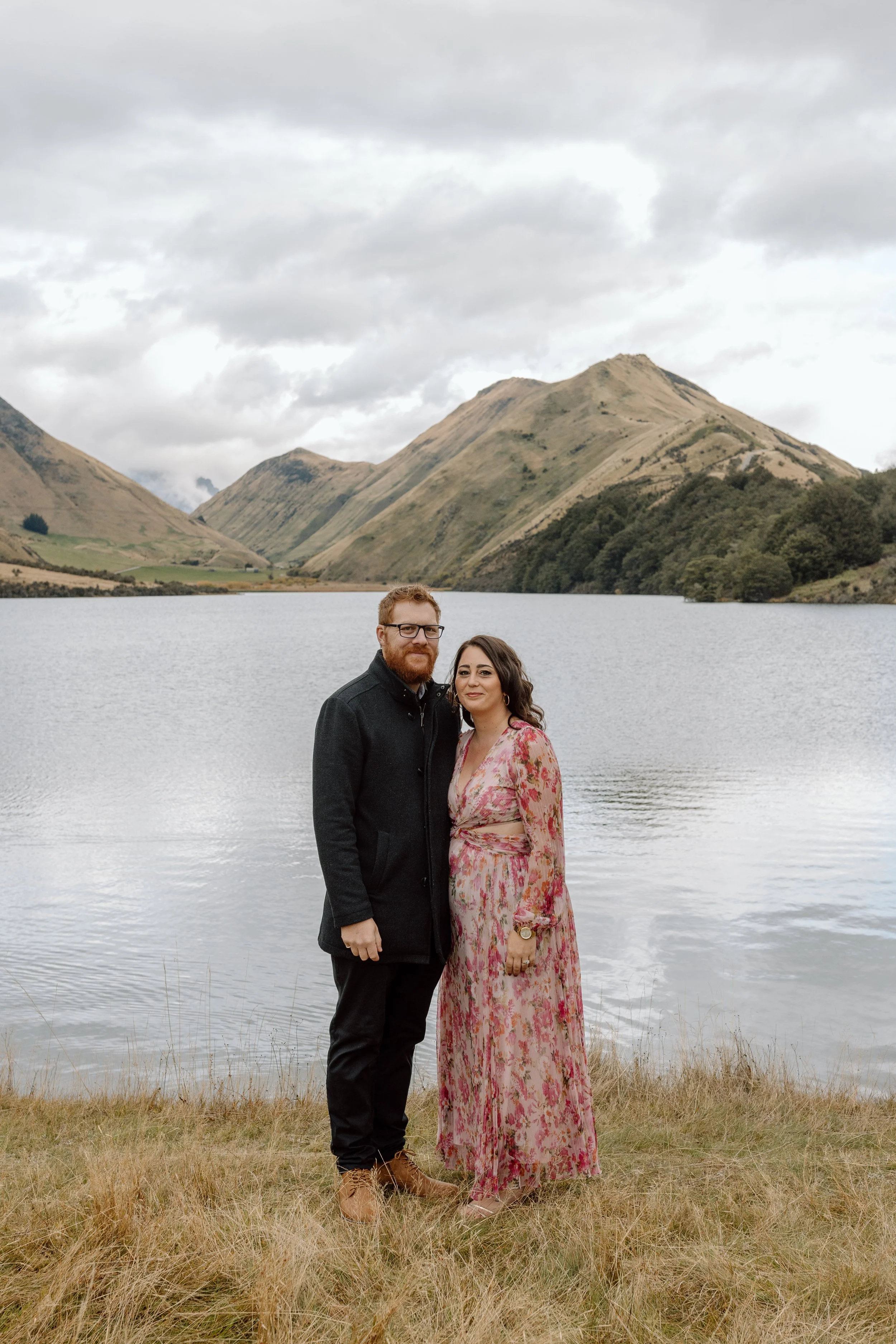 A man with glasses and a beard and a woman in a pink floral dress standing by a lake with mountains under cloudy skies in the background.