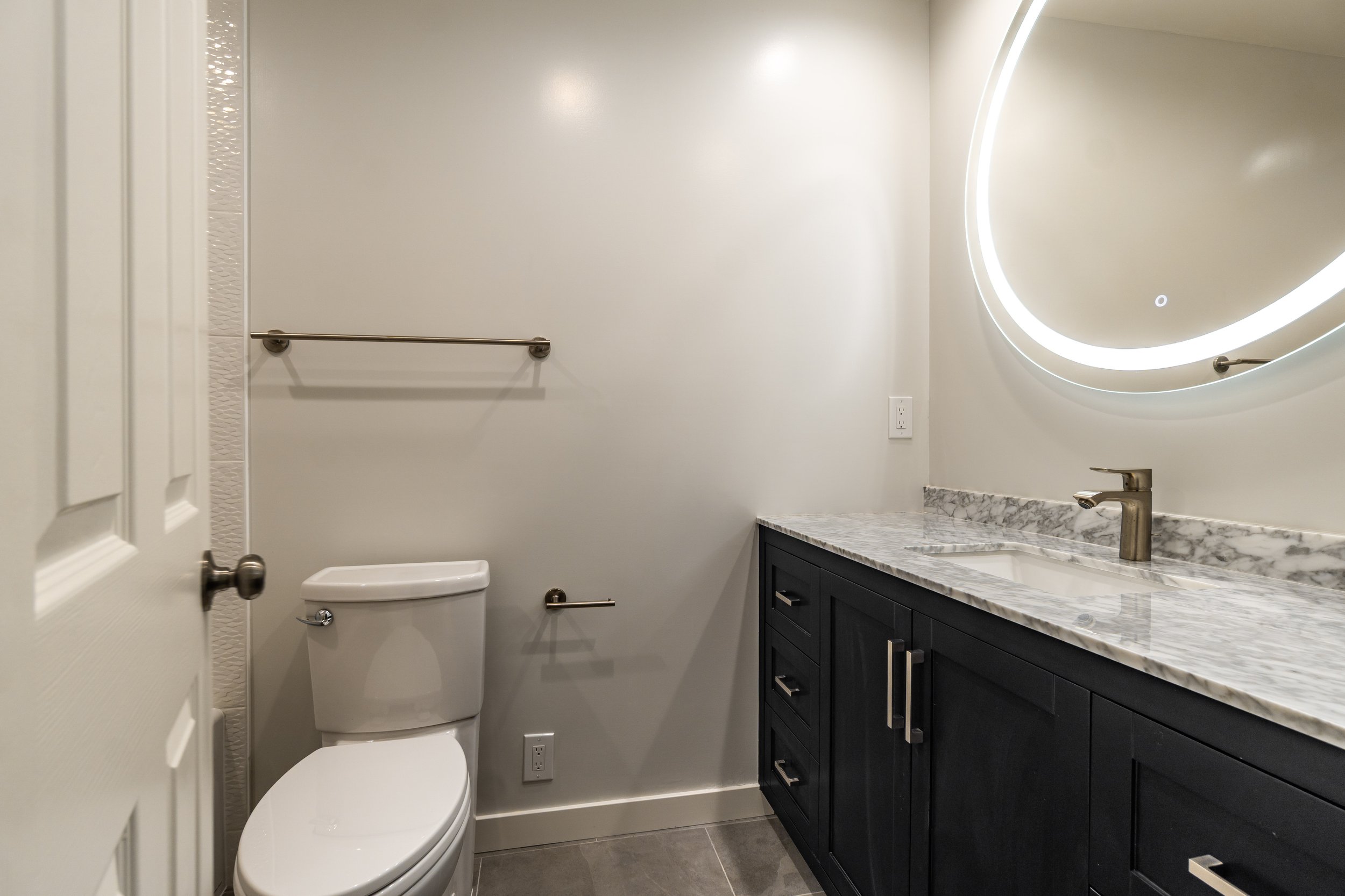 Modern powder bathroom with black vanity, marble countertop, circular LED mirror, and sleek fixtures.