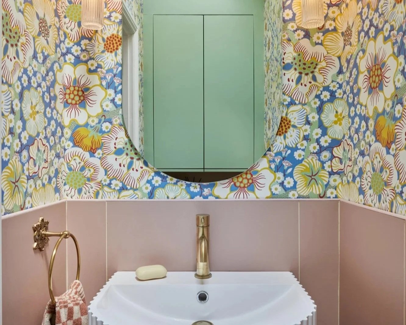 A bathroom with a floral patterned wallpaper featuring large yellow, white, blue, and red flowers. There is a round mirror above a white sink with a gold faucet. A soap bar is on the sink, and a gold towel ring with a pink and white checkered towel hangs beside it. A green cabinet is reflected in the mirror.