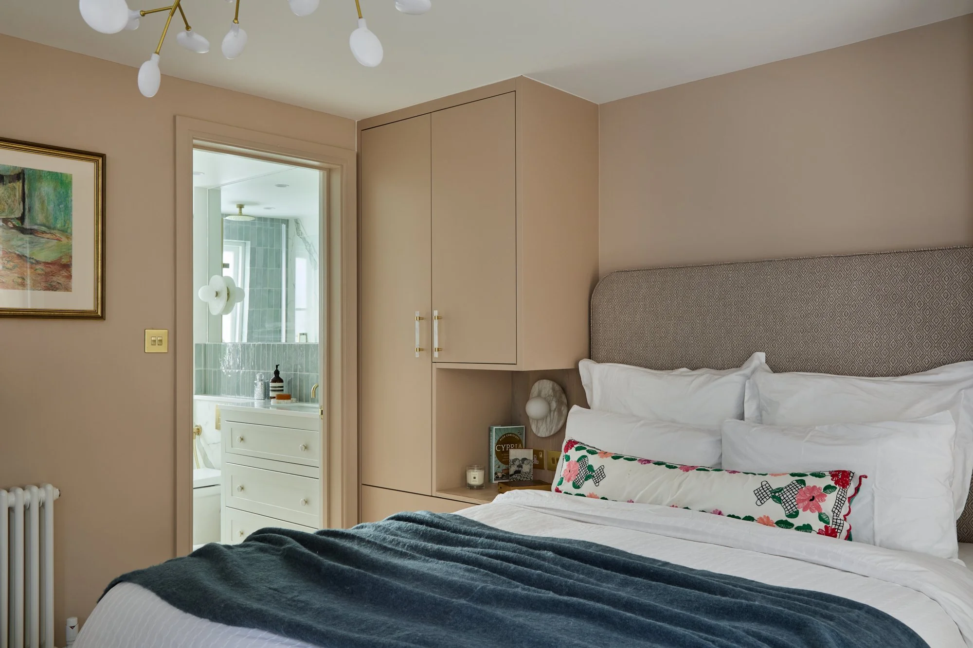A bedroom with a bed that has white bedding, a decorative pillow with a floral pattern, a dark blanket, and a headboard. There is a built-in cabinet above the bed, framed artwork on the wall, and a doorway leading to a bathroom with green-tiled walls and a vanity with a dark soap dispenser. The ceiling features a modern chandelier with white globes and brass accents.