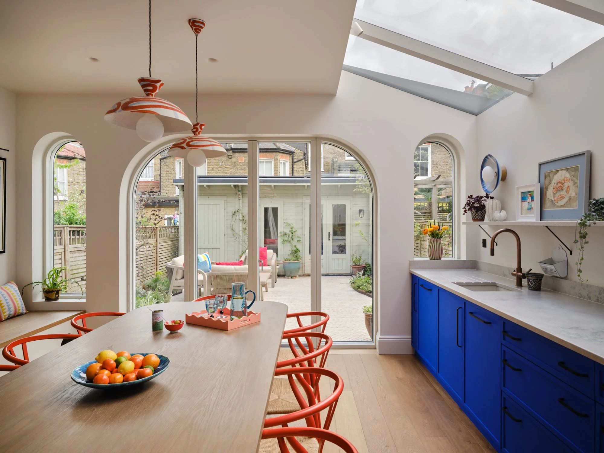 A bright kitchen with a wooden dining table, orange chairs, and a bowl of colorful fruits. The room has large arched windows and a sliding door leading to an outdoor patio. The kitchen features white walls, blue cabinets, and decorative art on the wall. A skylight provides natural light.