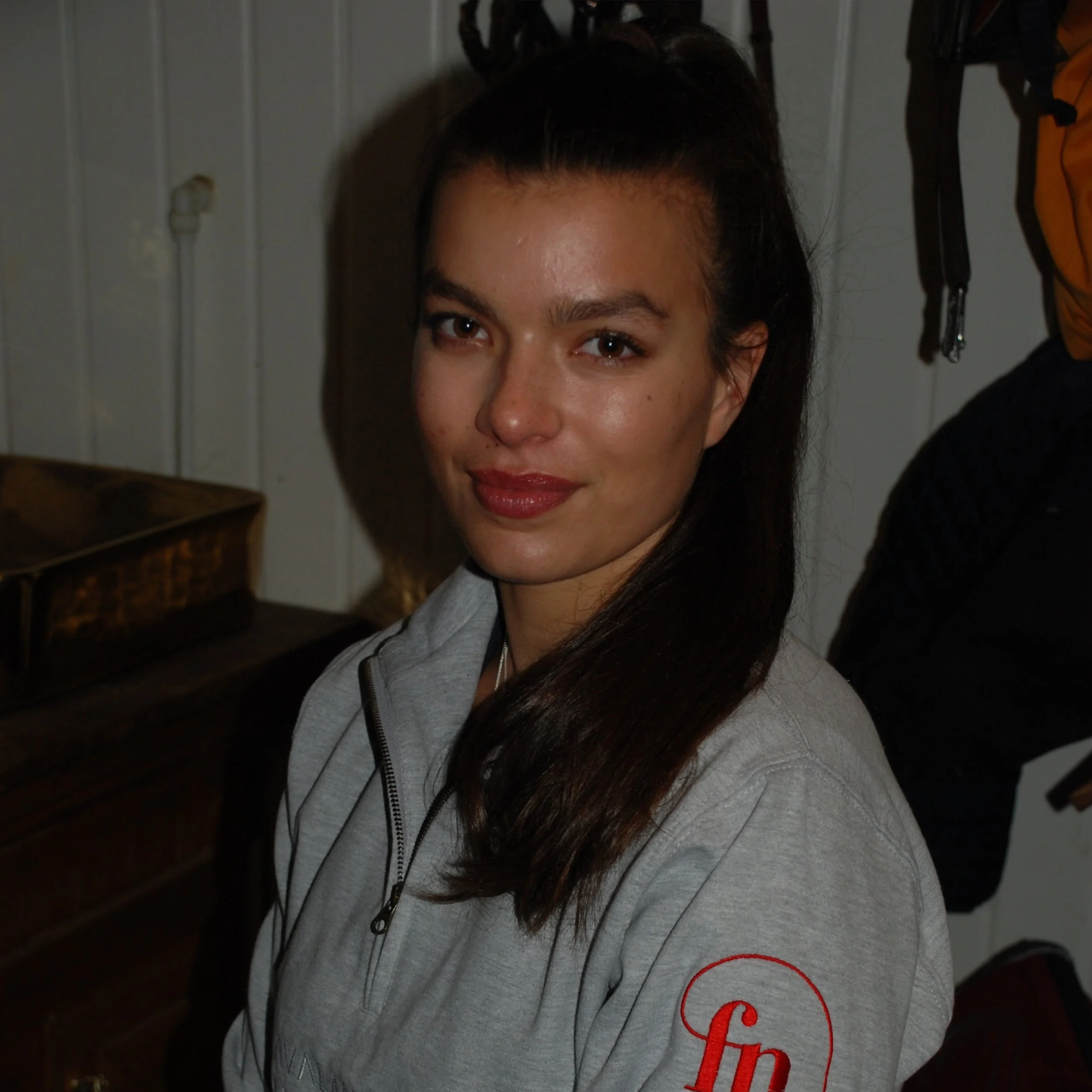 A woman with dark hair, wearing a light gray zip-up jacket with a red logo on the sleeve, sitting indoors near a wooden piece of furniture and a wall with electrical outlets.