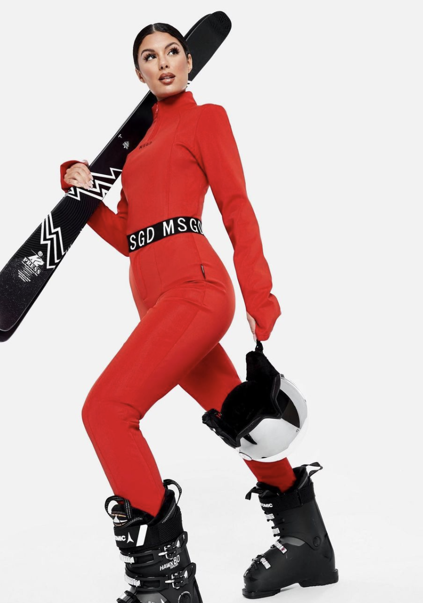 A woman dressed in a red ski suit holding a pair of skis over her shoulder and a white ski helmet in her other hand, standing on ski boots.
