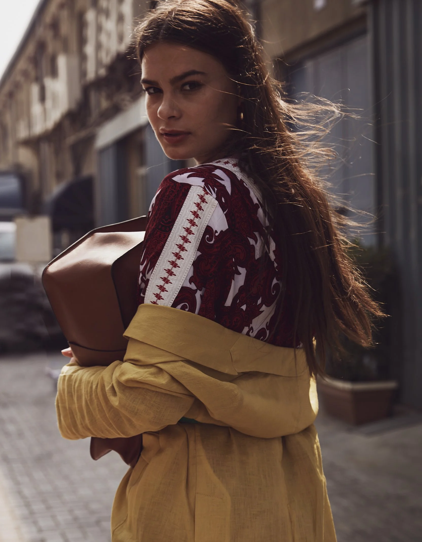 Young woman with long brown hair holding a coffee cup and wearing a yellow jacket and patterned top on a city street.