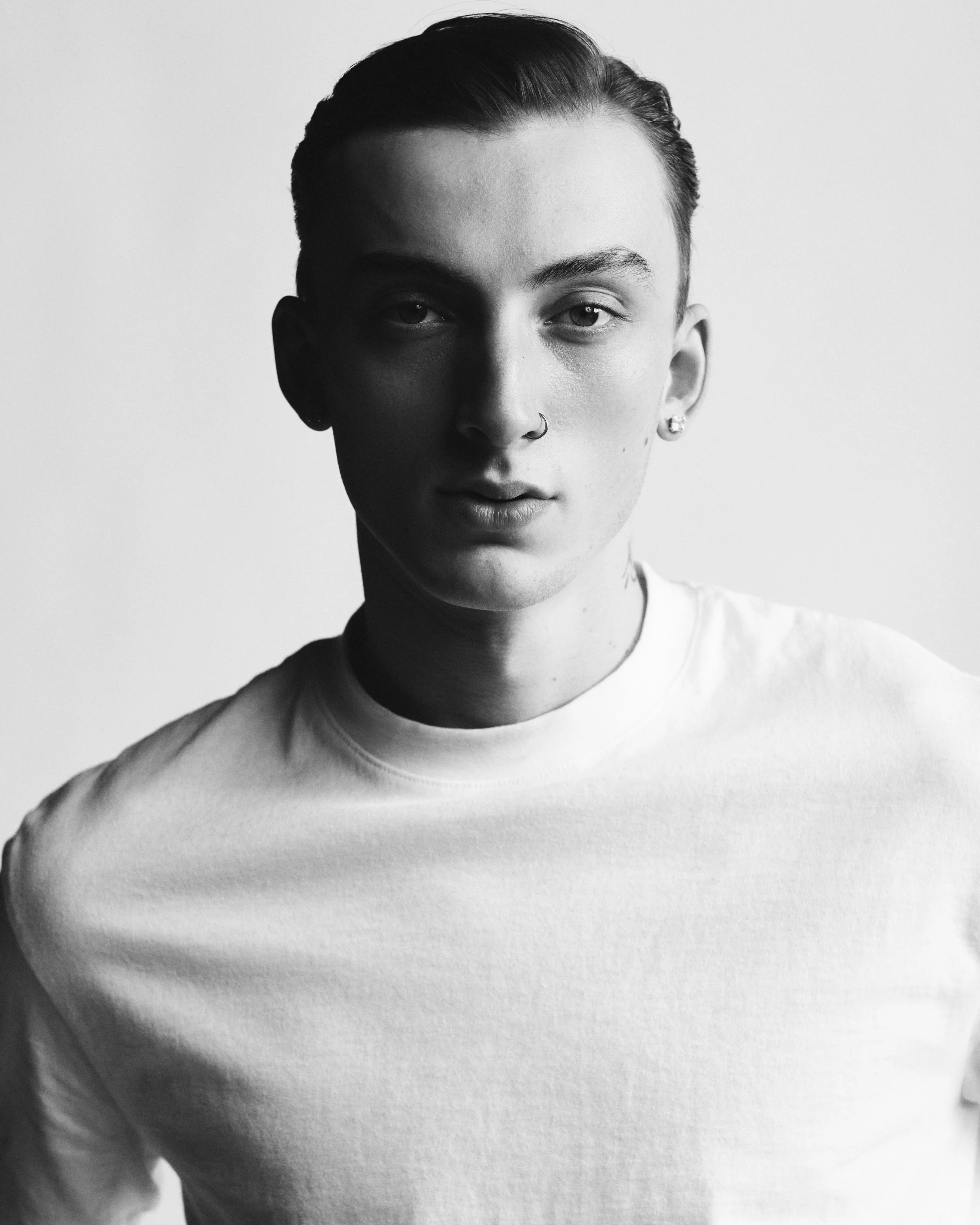 Black and white portrait of a young person with short hair, wearing a nose ring and earrings, dressed in a plain white t-shirt, looking directly at the camera.