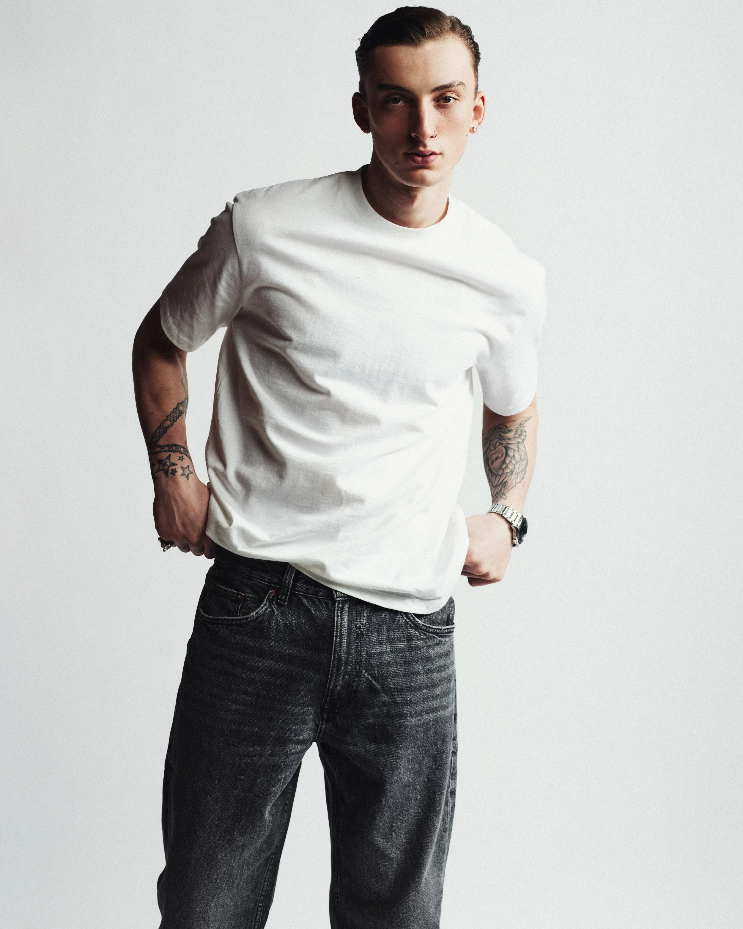 Young man with tattoos on arms wearing a white t-shirt and black jeans, standing against a gray background.