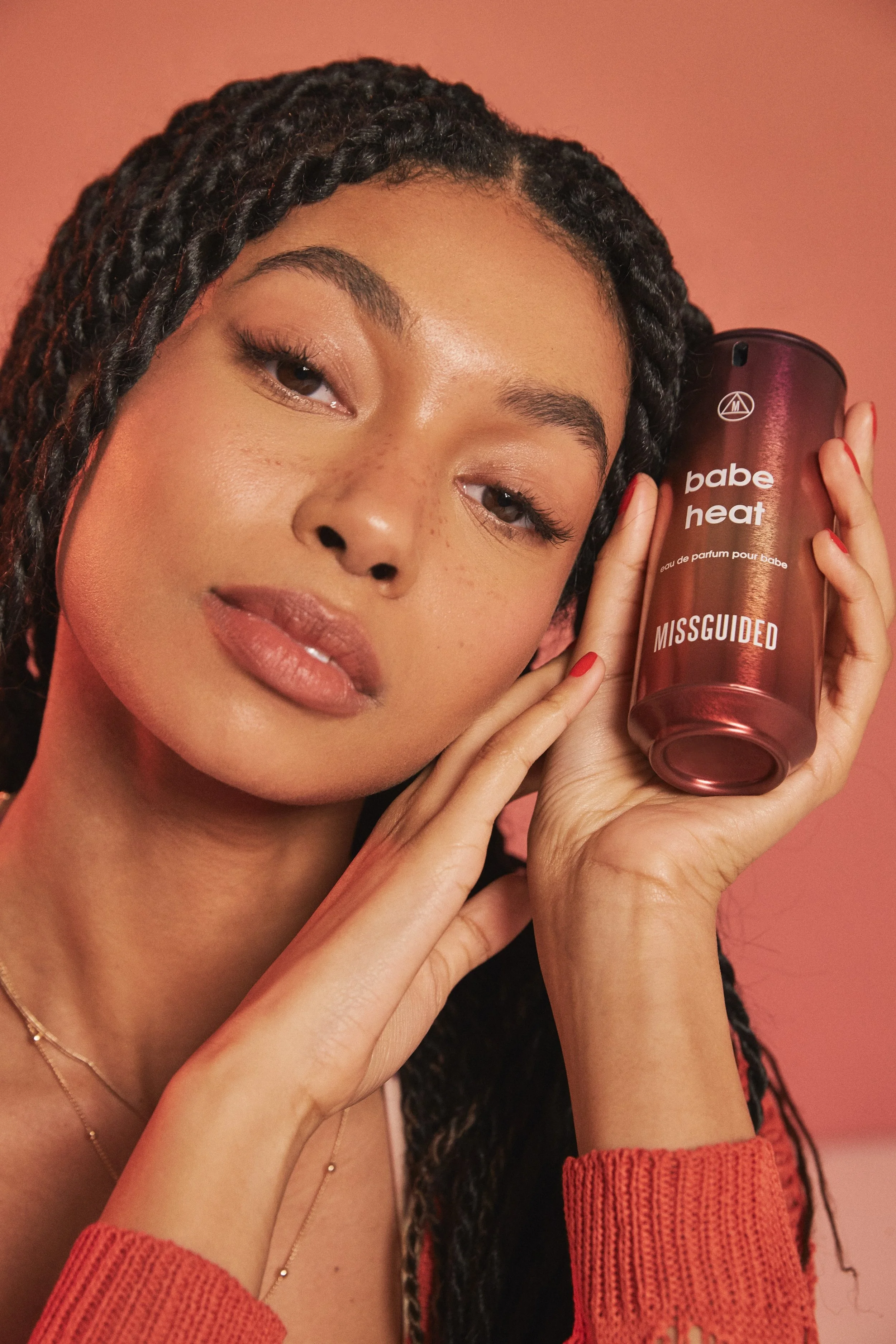 A young woman with braided hair holds a rose gold perfume bottle labeled 'babe heat' and 'MISGUIDED' close to her face, with a soft pink background.