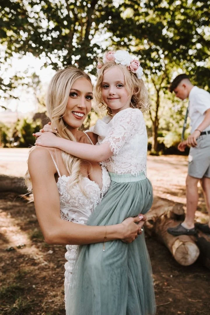 A woman holding a young girl in an outdoor setting with trees in the background. The woman is smiling and the girl is wearing a floral crown and smiling. A boy in a white shirt and shorts is in the background.