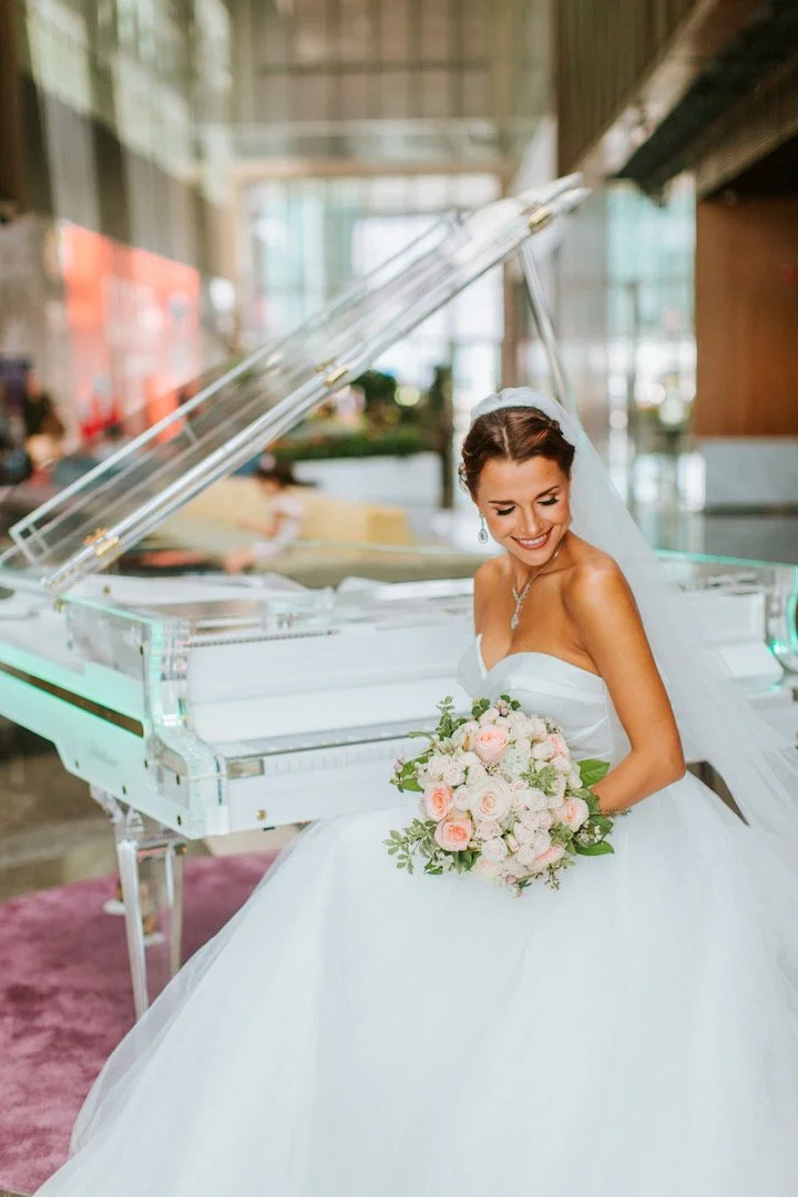 A bride in a white wedding gown sitting at a white grand piano, holding a bouquet of pink and white flowers, smiling and looking down in a modern, well-lit indoor venue.