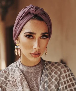 A woman with a purple headscarf, makeup, and earrings, wearing patterned clothing, indoors.
