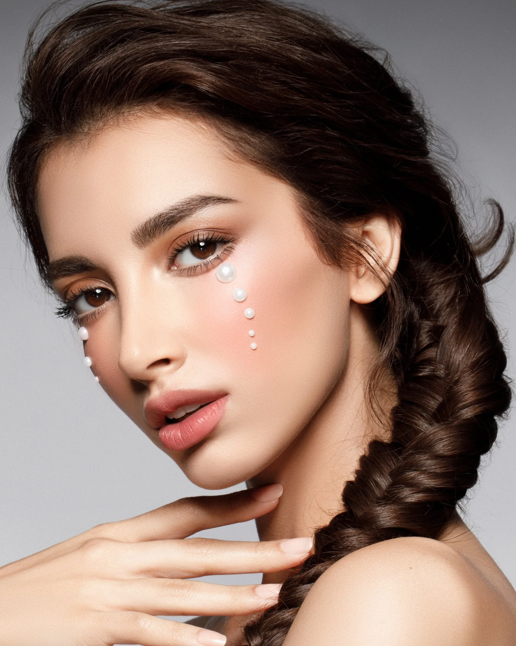 A woman with long brown hair styled in a braid, makeup, and decorative pearls on her face.