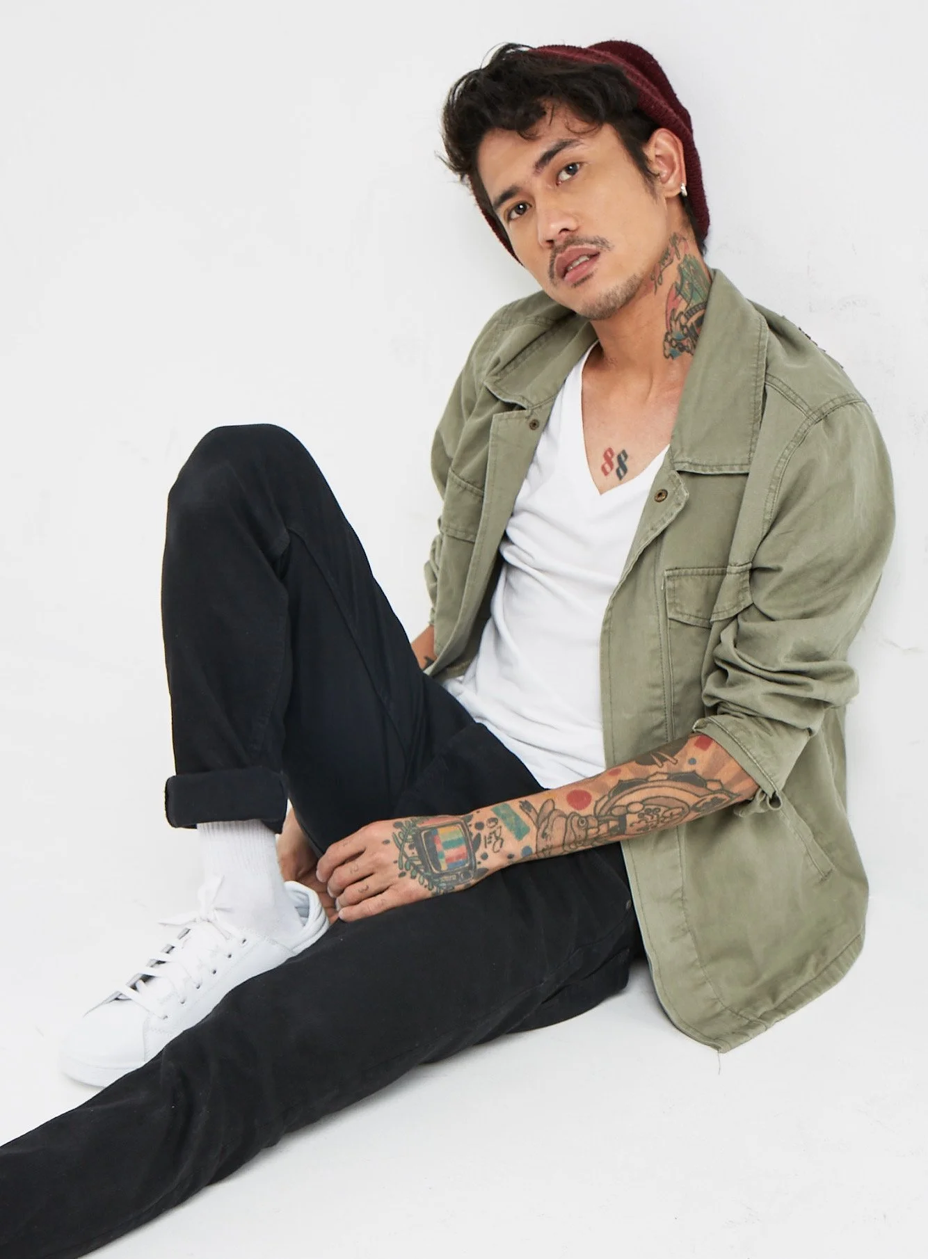 A young man sitting on the floor against a white background, wearing a khaki jacket, white t-shirt, black pants, white sneakers, and a burgundy beanie. He has tattoos on his arms, neck, and chest, and is looking at the camera with a serious expressio