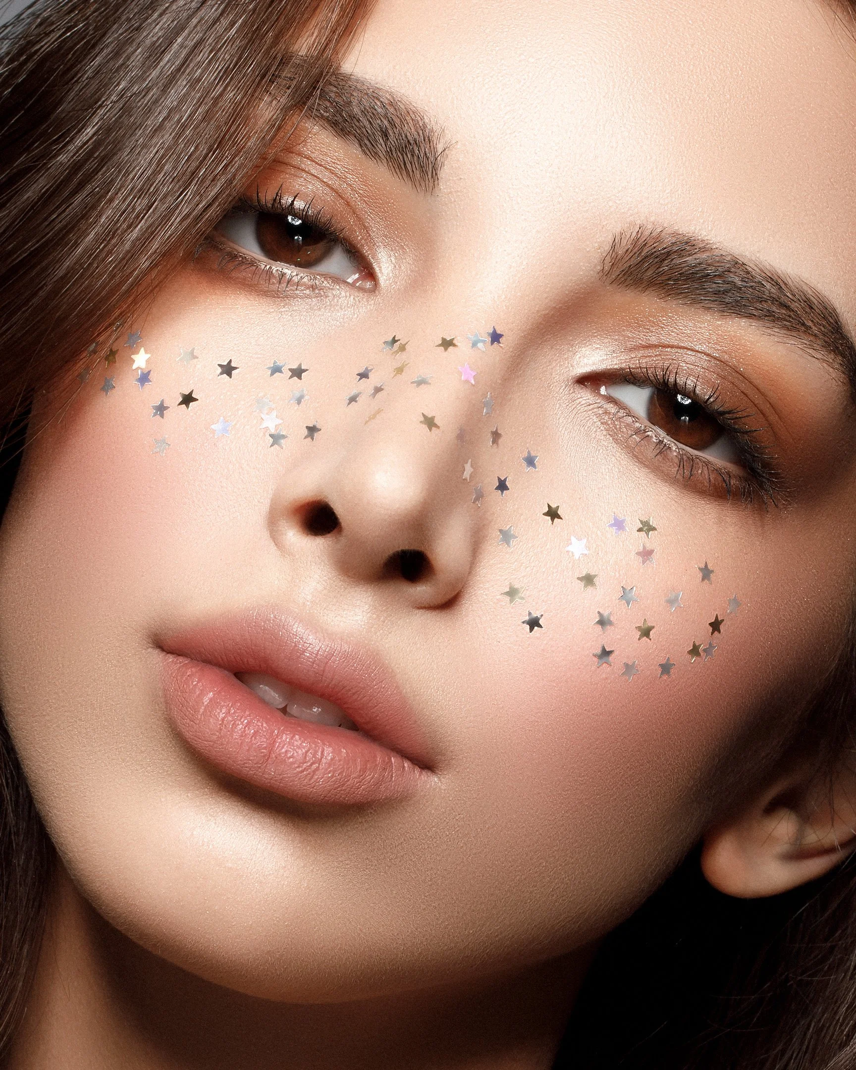 Close-up of a woman's face with star-shaped confetti on her cheek, natural makeup, brown eyes, and long brown hair.