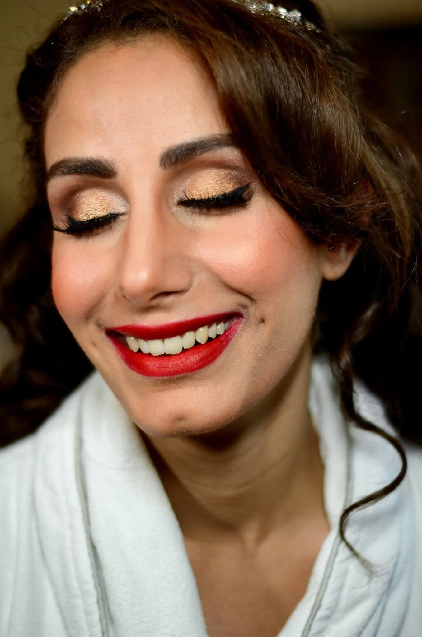 A woman with closed eyes, smiling, showing her teeth, wearing makeup with gold eyeshadow, black eyeliner, and red lipstick.