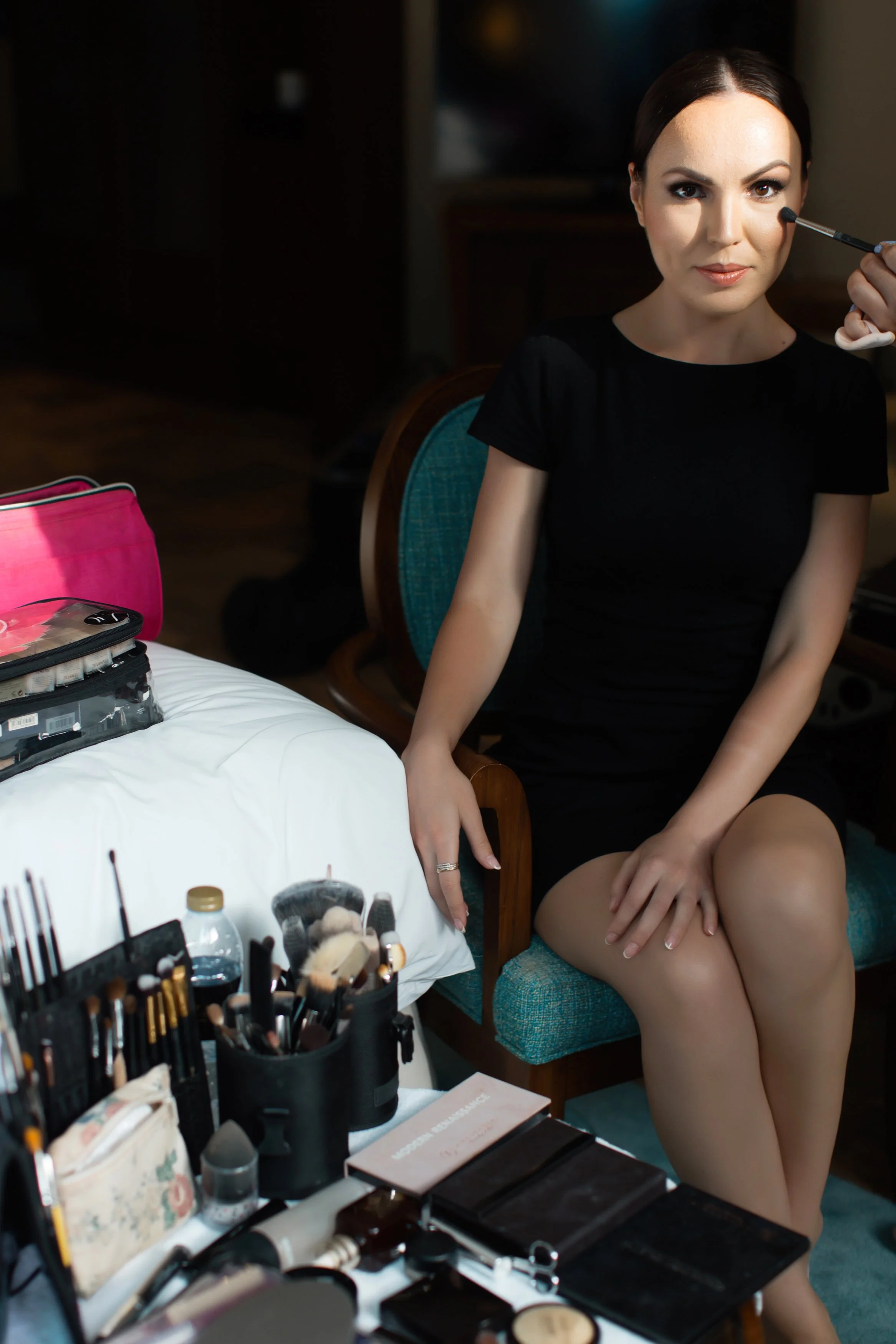 Woman sitting in a chair with makeup tools and products on a table beside her, applying makeup with a brush.