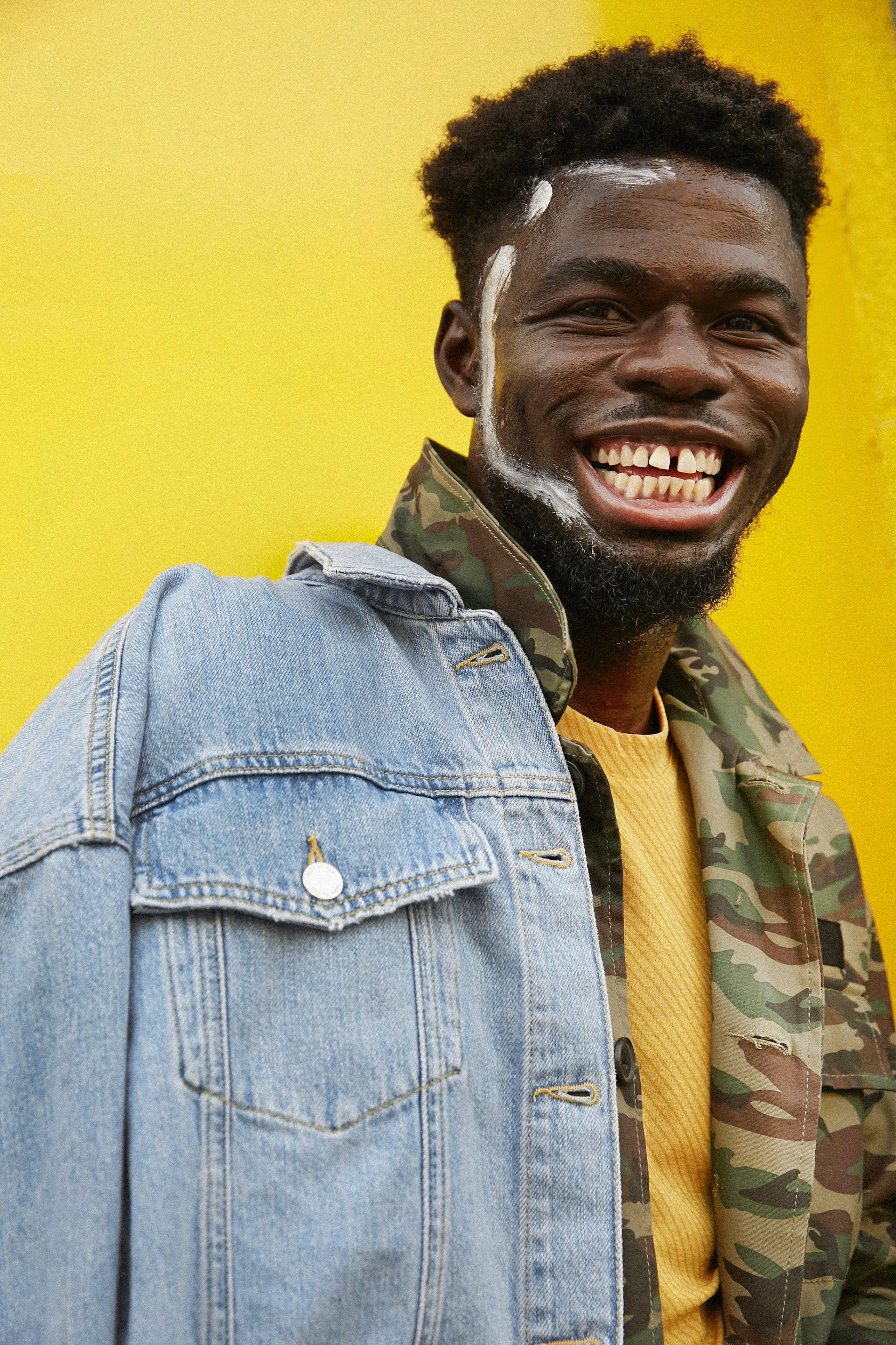 Smiling young man with a beaded necklace wearing a denim jacket, yellow shirt, and a camouflage-patterned coat standing against a yellow background.