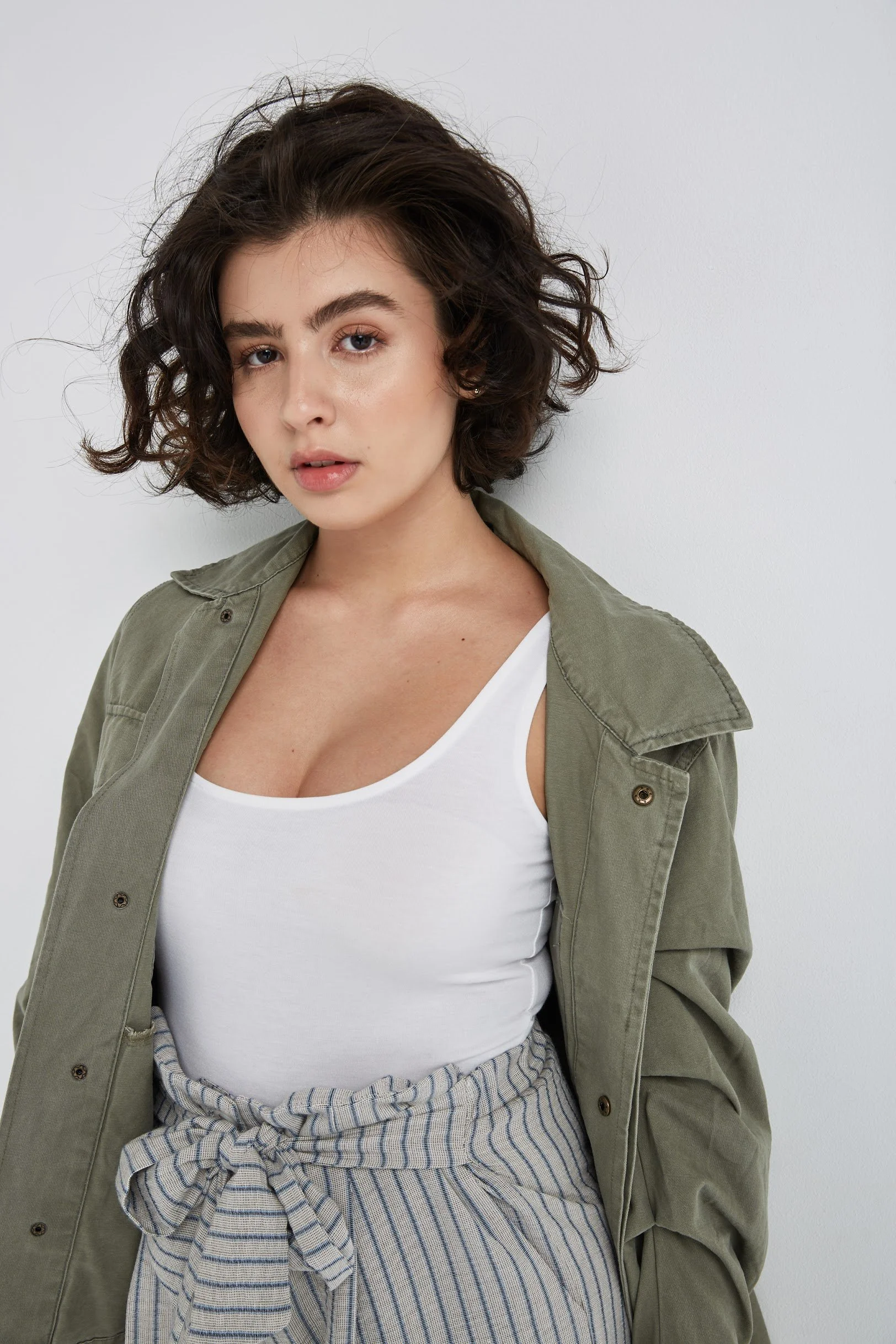 A young woman with short curly dark hair wearing a white tank top, olive green jacket, and striped high-waisted pants, standing against a plain white background.