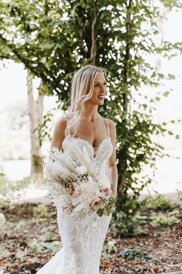 A bride in a white lace wedding dress holding a bouquet of pale pink roses and white flowers outdoors surrounded by green trees.