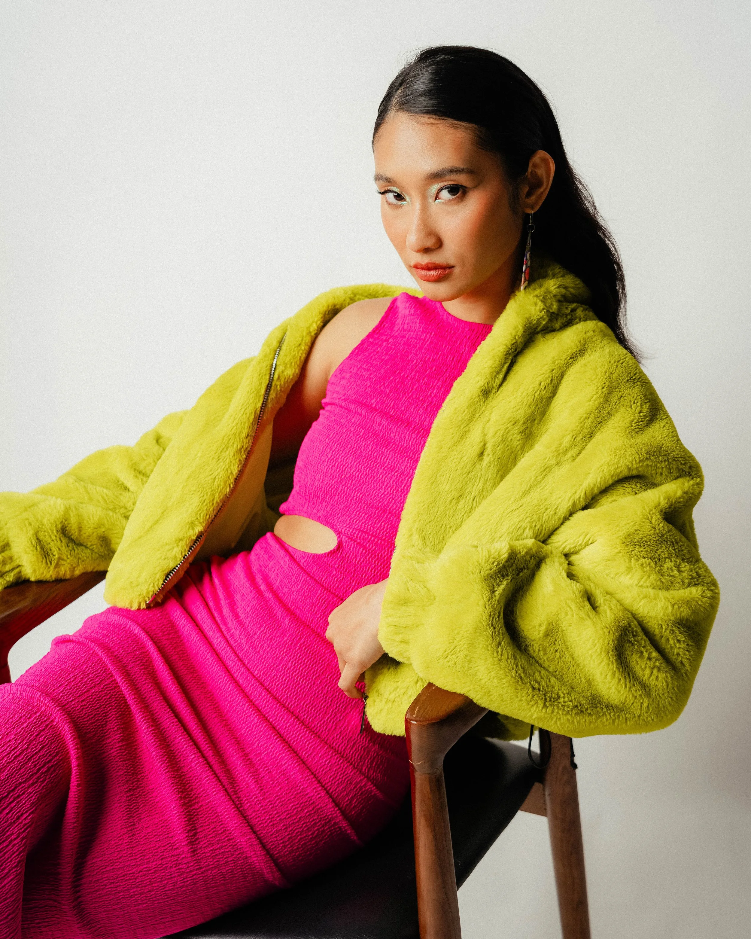 A young woman with dark hair and makeup, wearing a bright pink dress with a cutout, and a yellow furry jacket, sitting on a wooden chair against a plain white background.