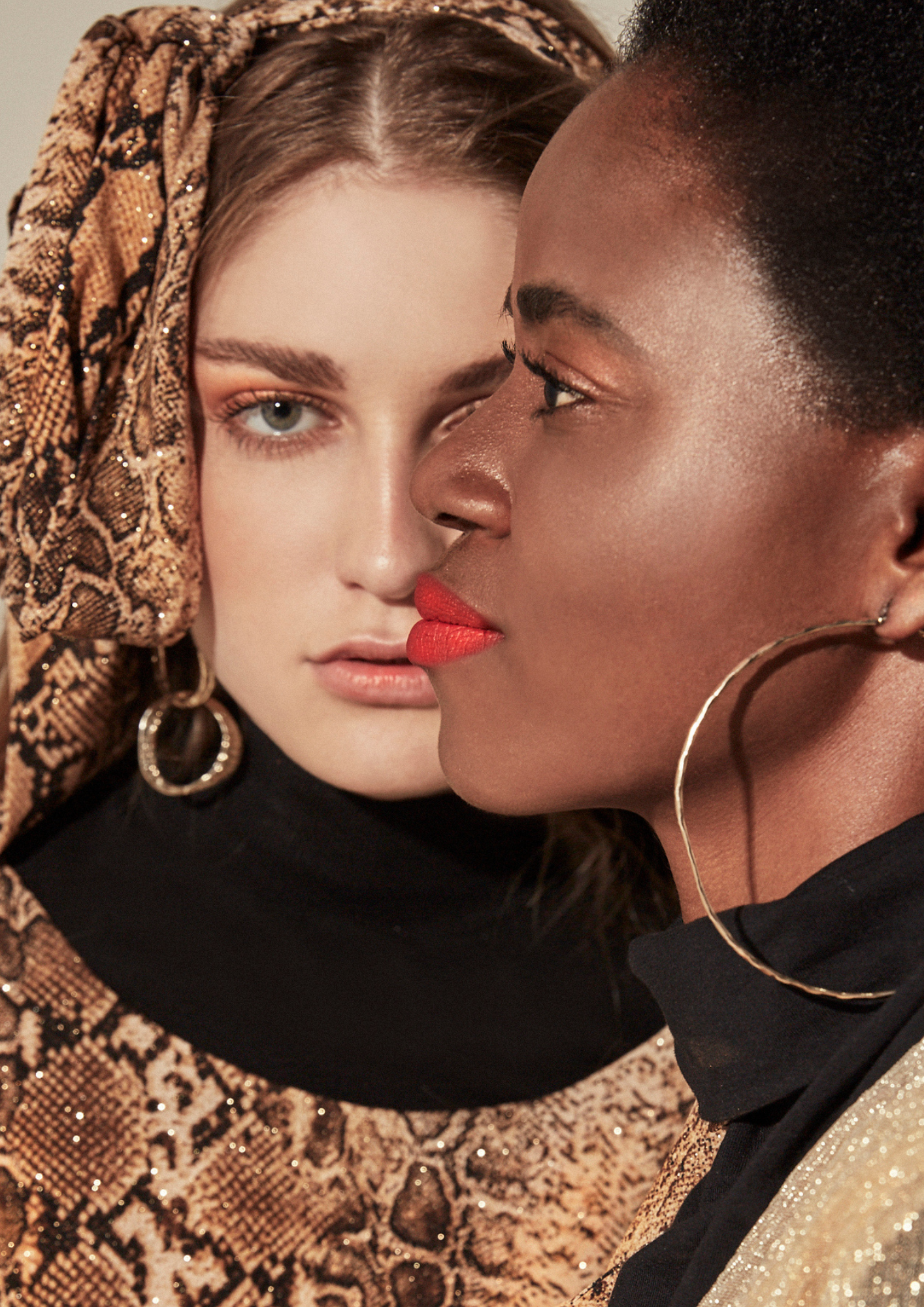 Close-up portrait of two women, one with fair skin and blue eyes wearing a snake-print headscarf, and the other with dark skin and short hair wearing large hoop earrings. They are positioned closely, with the woman on the left looking at the camera w