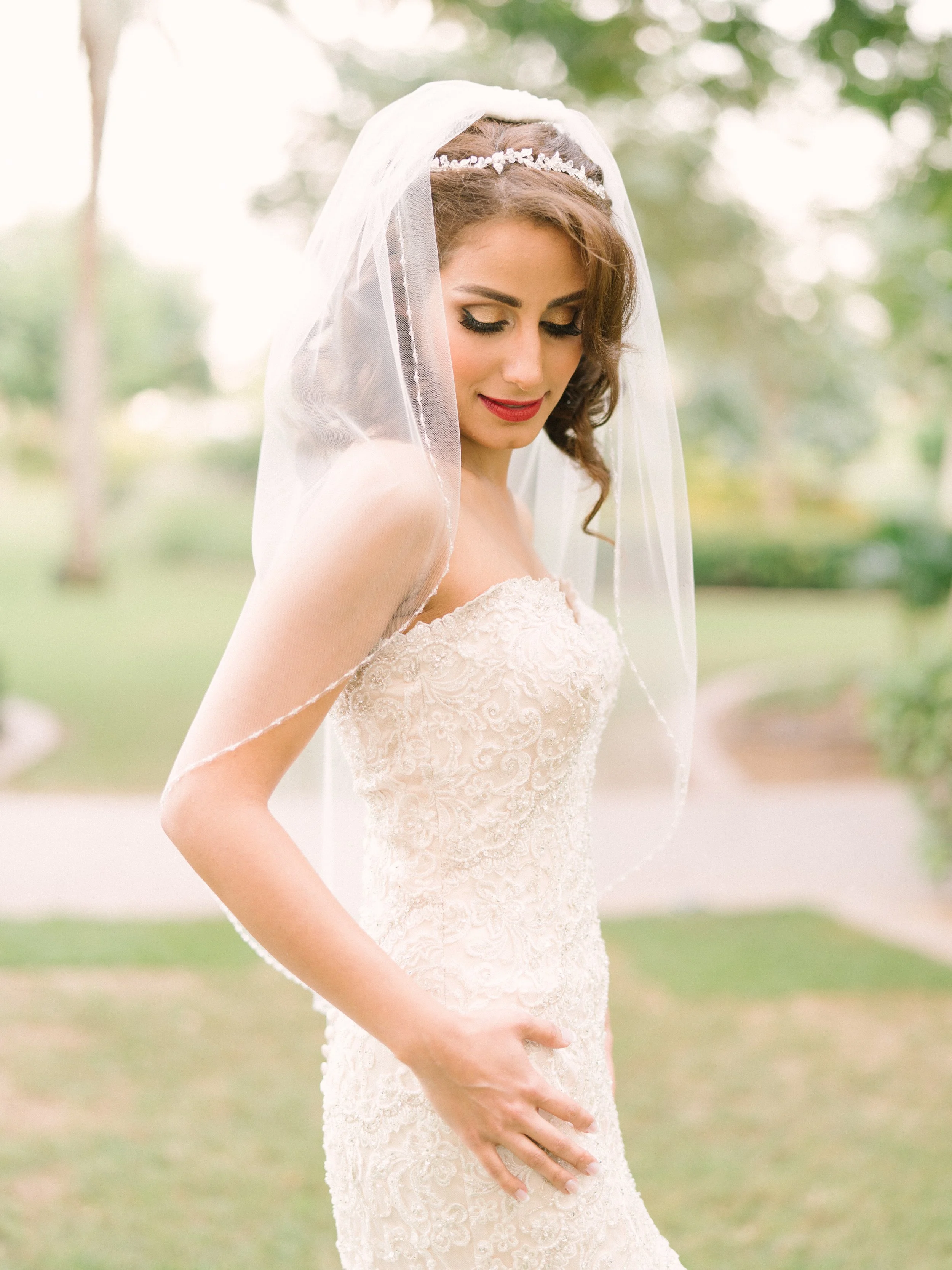 A bride in a lace wedding gown with a veil and tiara, outdoors in a park-like setting.